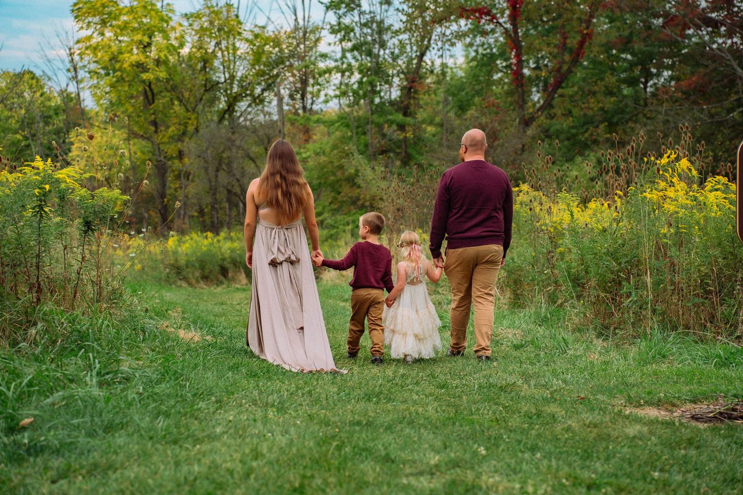 Family of four walks hand-in-hand through lush green meadow with wildflowers in autumn.