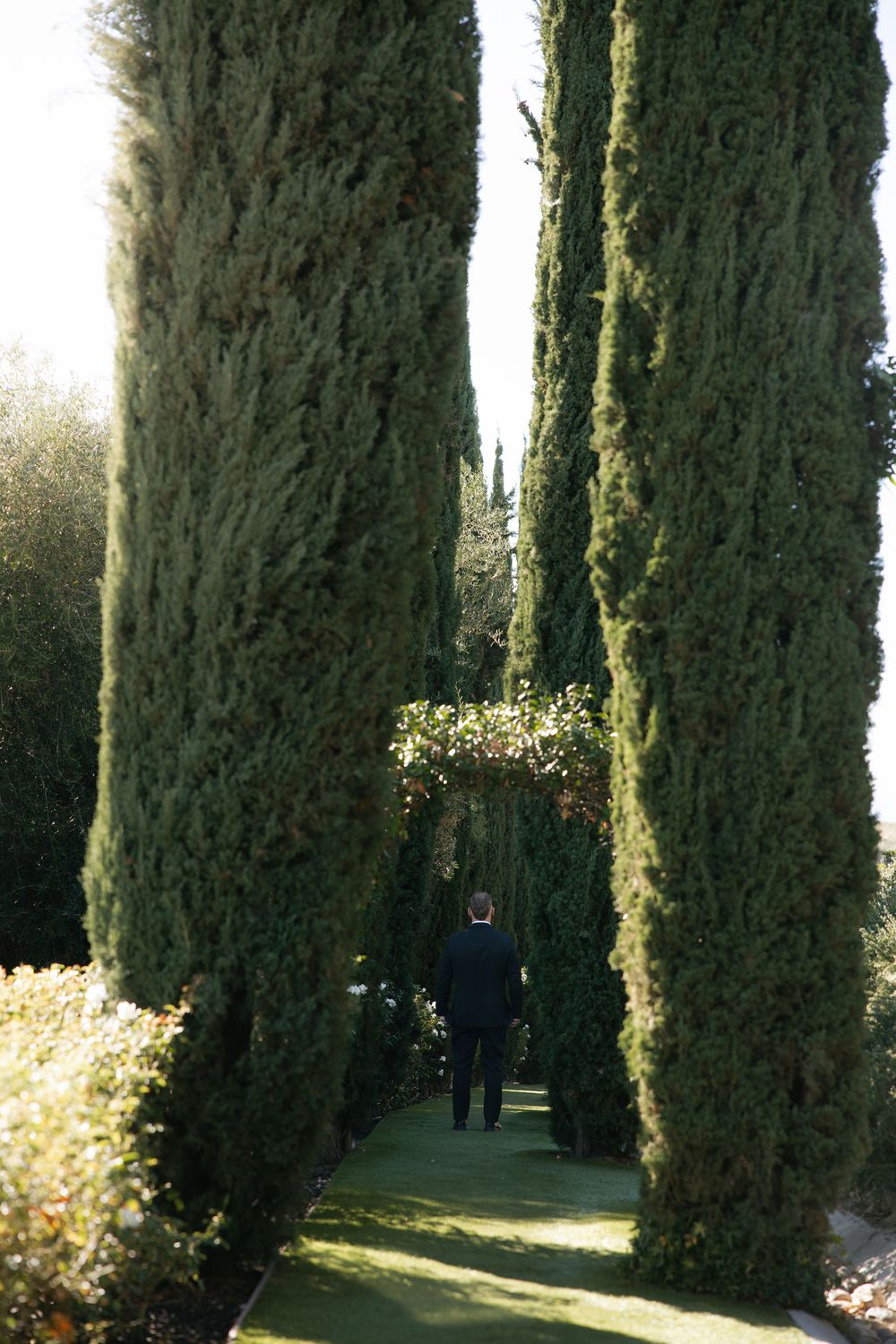 Groom walks through tall cypress tree lined garden path at wedding venue.