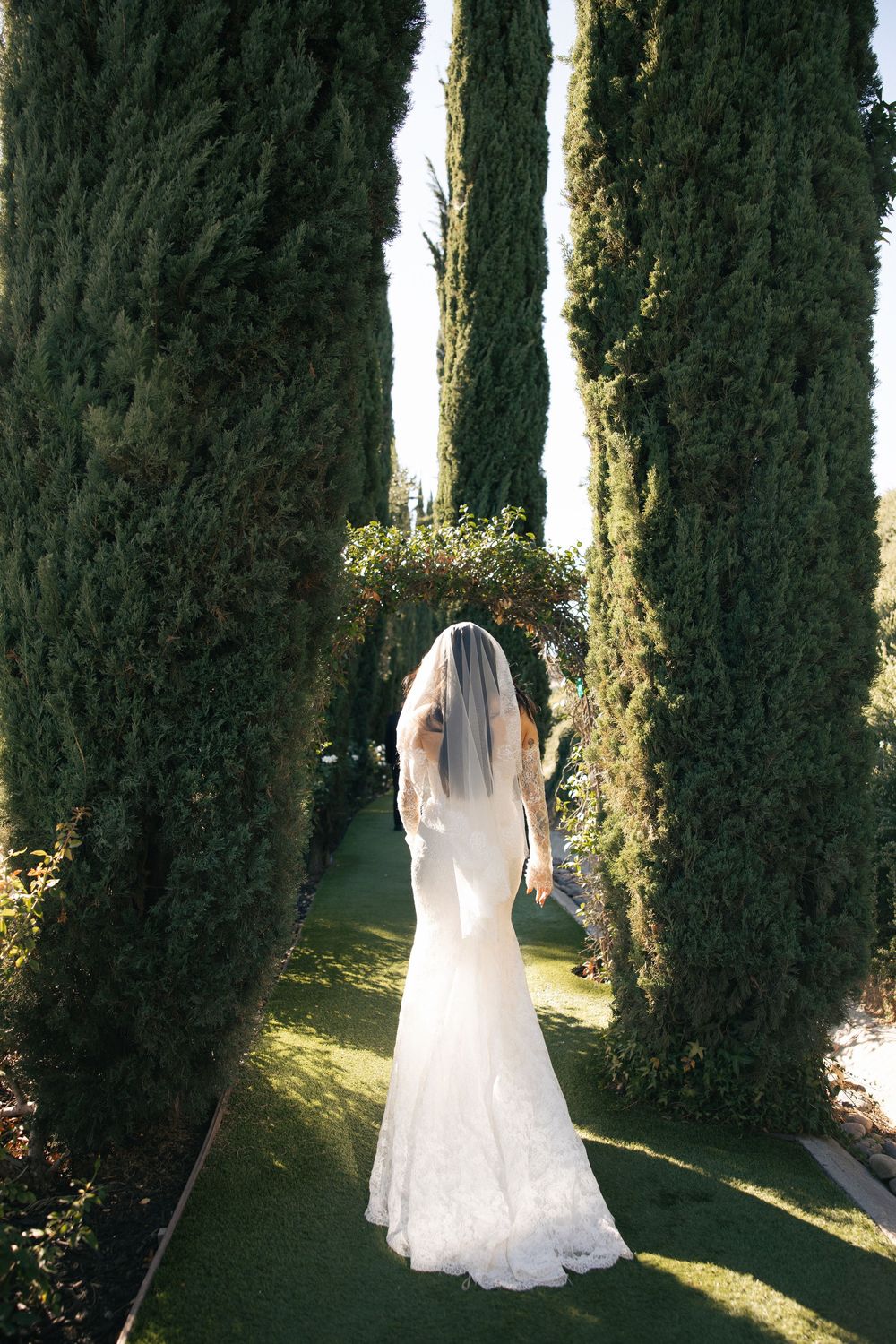 A bride in a lace wedding gown and veil walks between a tree-lined path at outdoor wedding venue in SoCal.