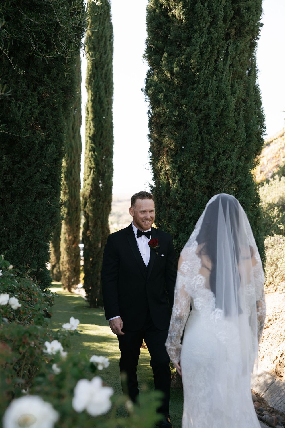 Groom in black tuxedo smiles at bride in lace gown and veil after first look along a tree-lined path at outdoor wedding venue in SoCal.