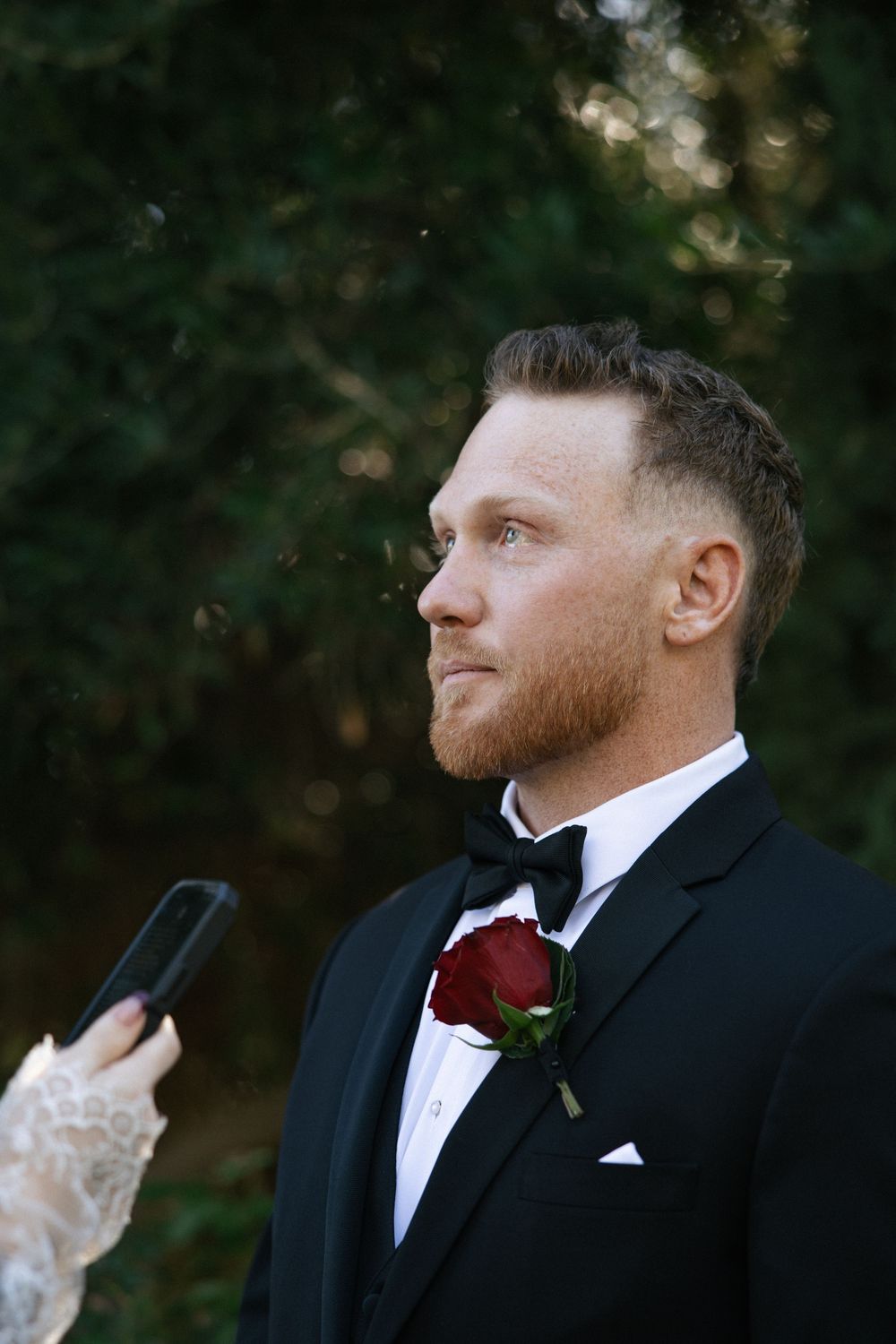 Groom in black tuxedo with red rose boutonniere during outdoor wedding ceremony, getting emotional