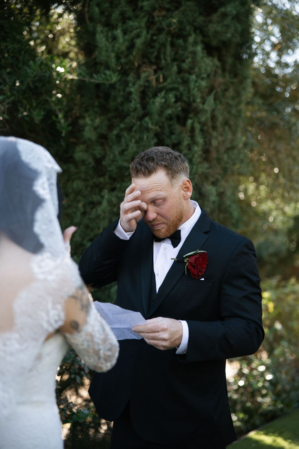 Emotional groom wiping tears while reading vows at outdoor wedding ceremony, wearing black tuxedo with red rose boutonniere.