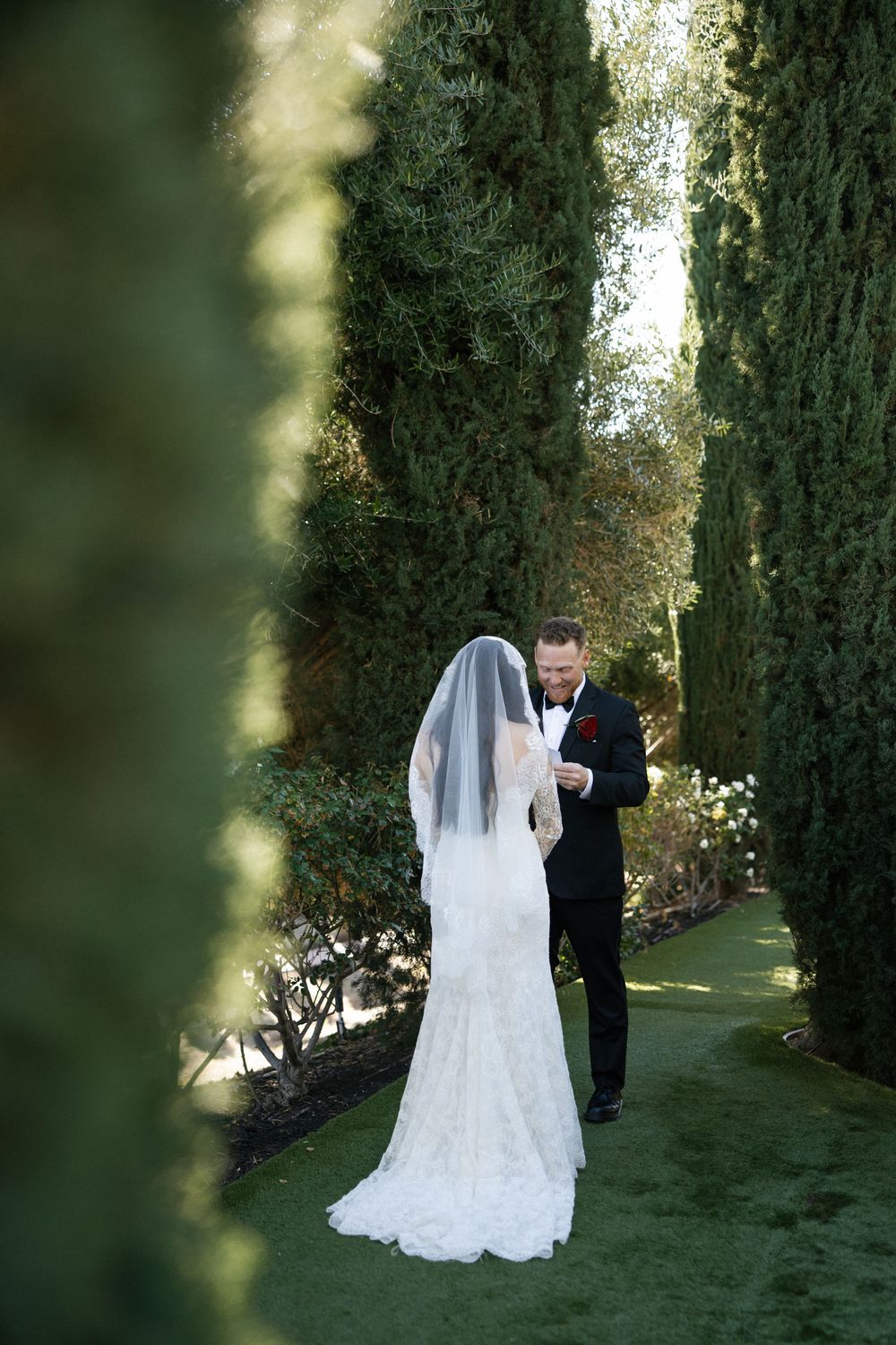 A bride and groom share an intimate moment on a garden path lined with tall cypress trees sharing their private vow