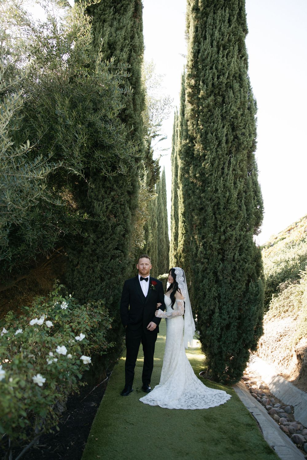 Bride and groom stand among tall cypress trees on a lush garden path, bride in lace gown with long veil.