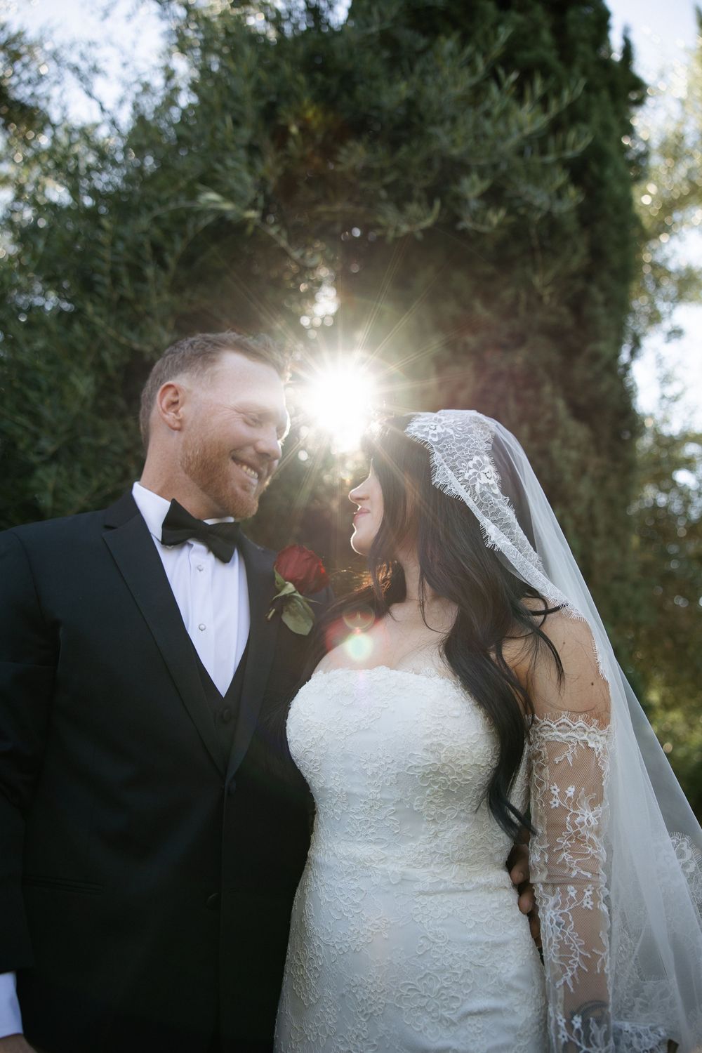 Bride and groom touching foreheads with sun flare between them, wearing wedding dress with veil and black tuxedo outdoors.