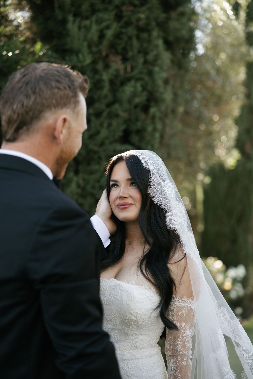 Bride in lace wedding dress and veil smiles at groom during outdoor wedding ceremony.