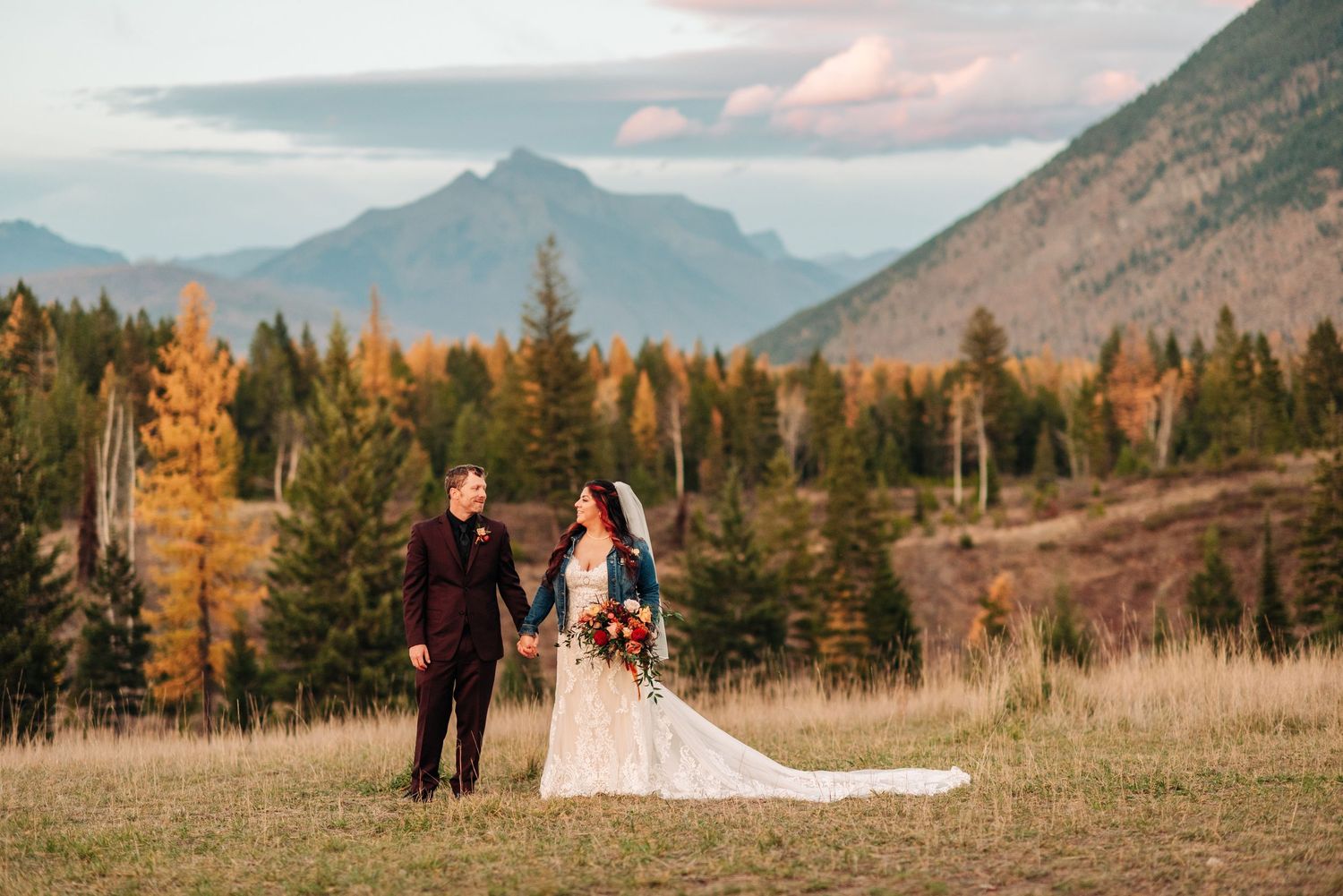 Bride and groom holding hands in a mountain meadow with autumn trees and peaks in the background.