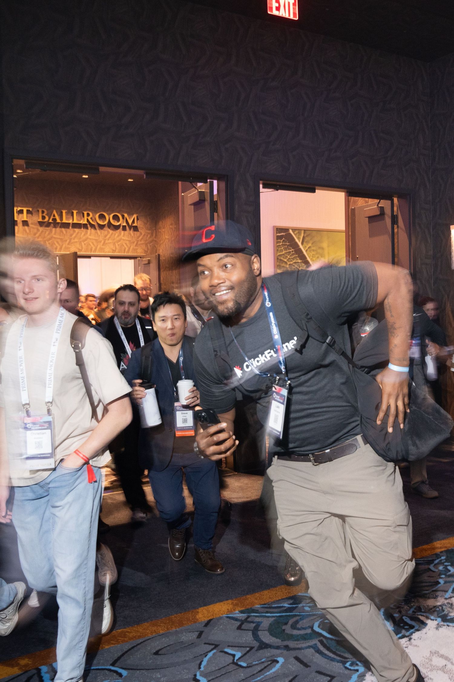 Man in black cap and t-shirt poses energetically at a ballroom event surrounded by a crowd of attendees.