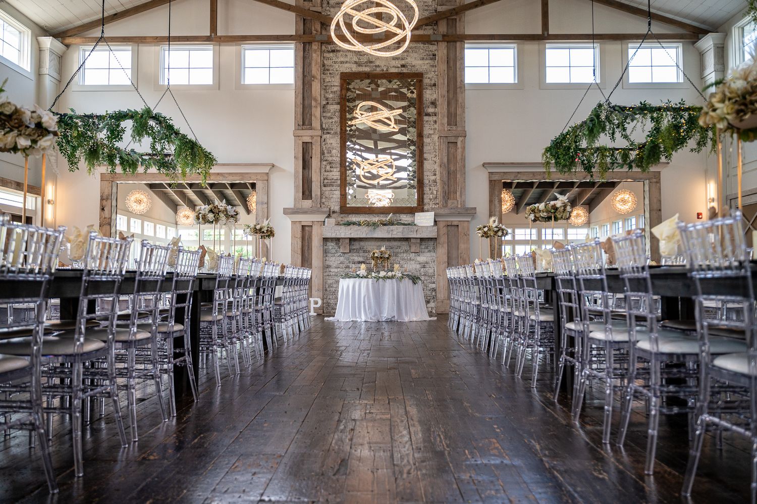Elegant wedding reception hall with long tables, ghost chairs, hanging greenery installations, and a vaulted ceiling with chandelier.