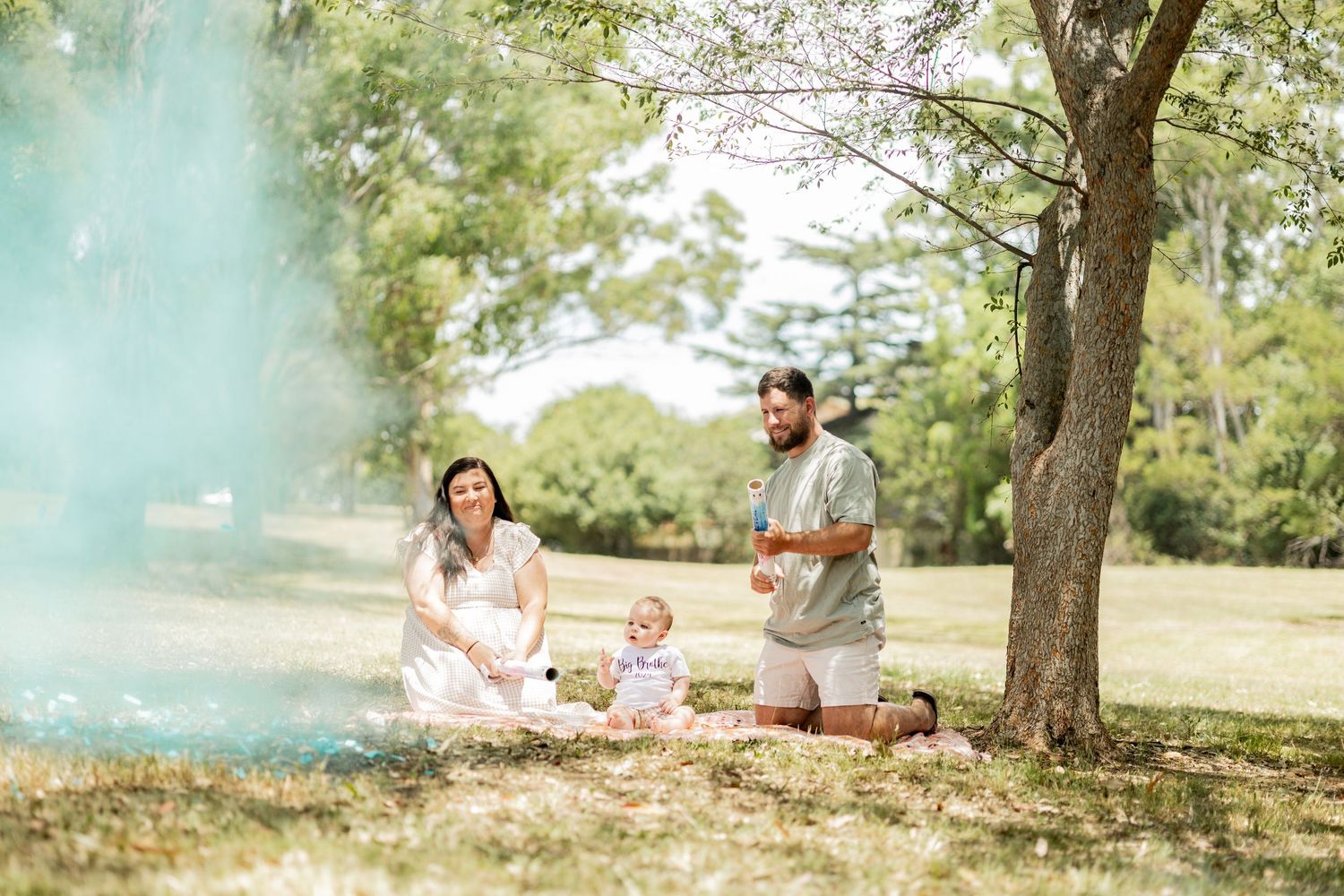 Family gender reveal with teal smoke in sunny park setting.