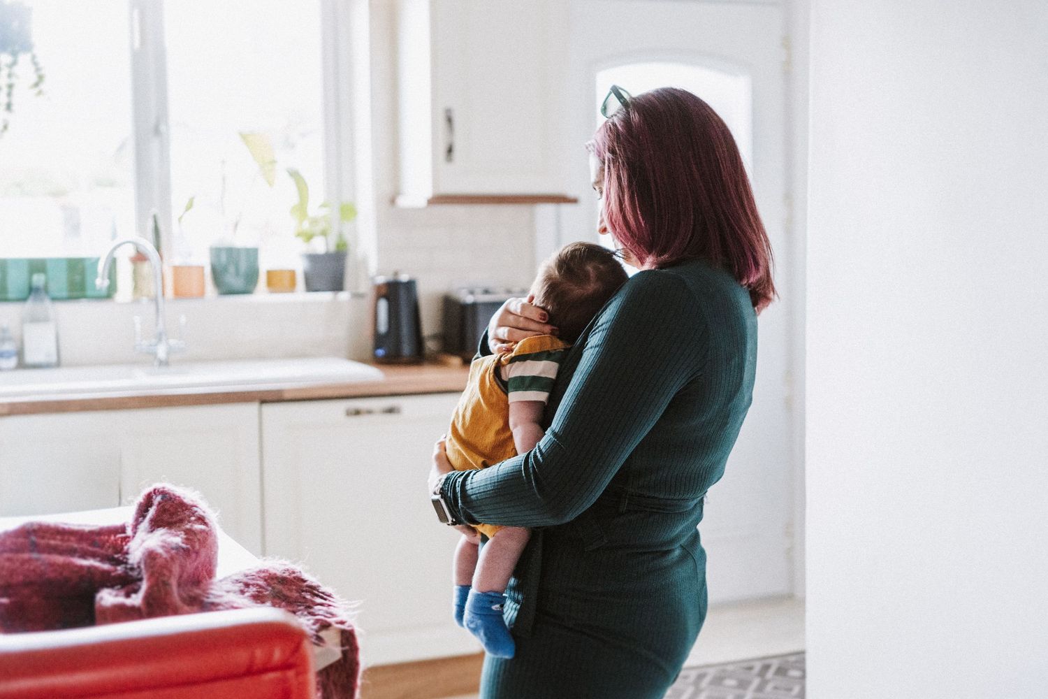 Person in dark green sweater holds and feeds a baby while standing in a bright modern kitchen interior.