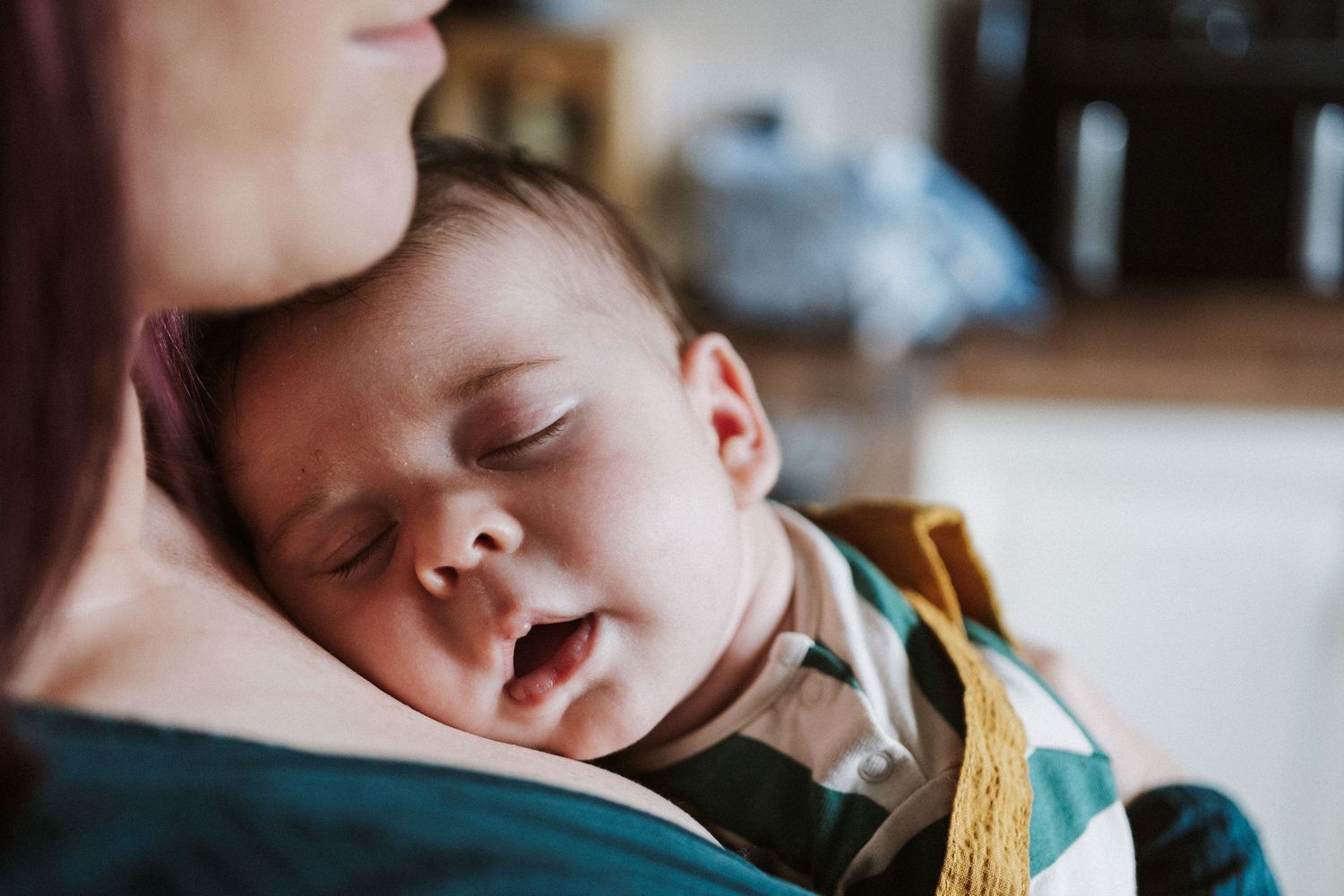 A sleeping baby rests peacefully against someone's shoulder in a warm, natural light setting.