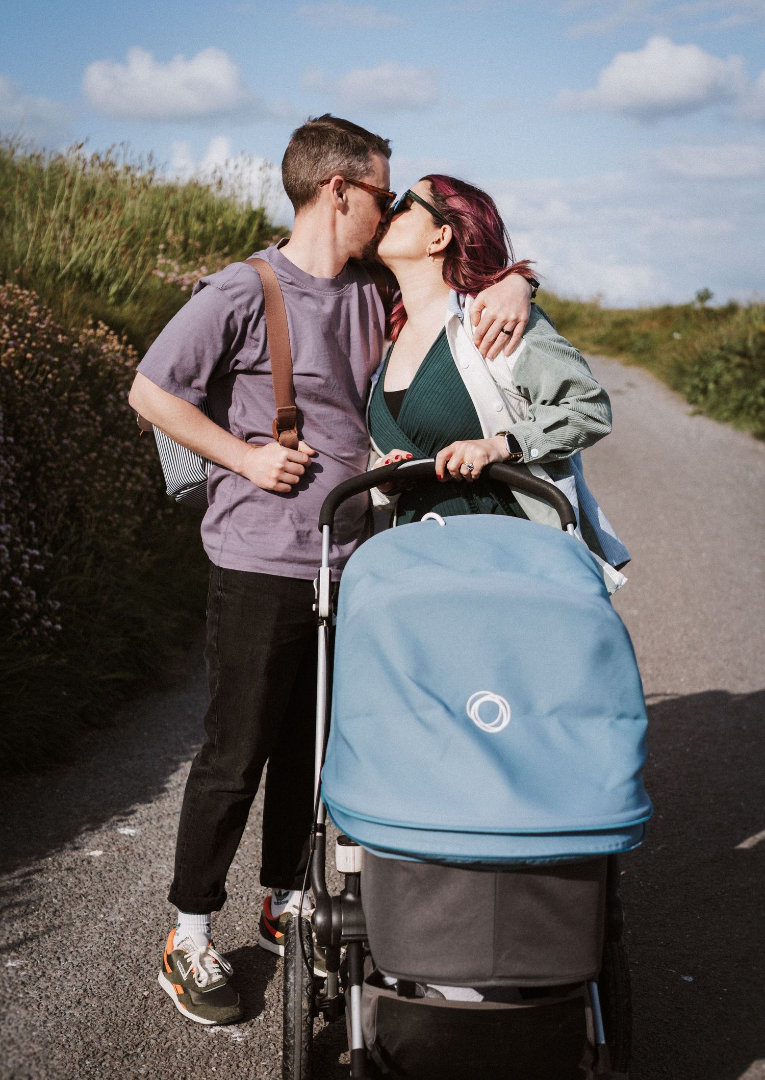 A couple shares an intimate moment on a path while standing next to a blue and gray baby stroller.