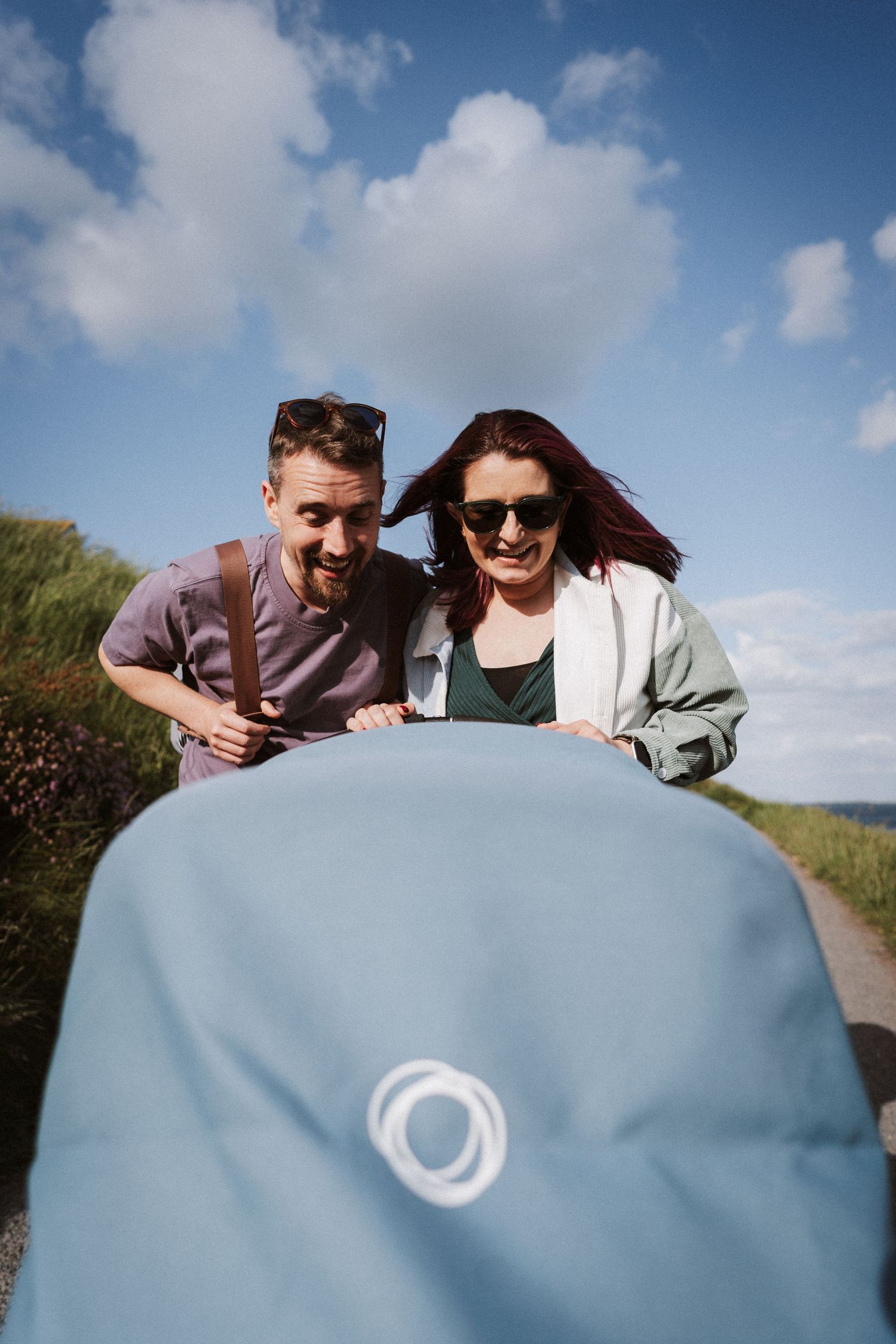 Two people peek playfully over a blue surface against a bright sunny sky with white clouds.