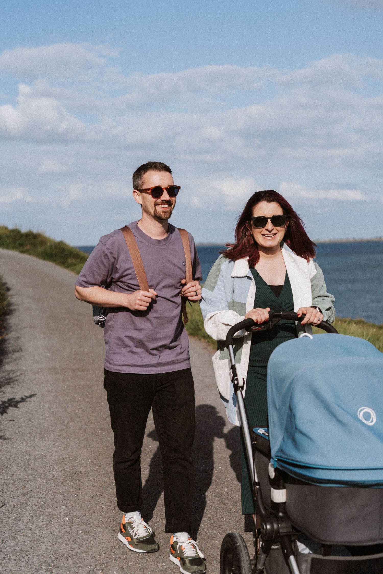 A couple walks along a coastal path on a sunny day, pushing a baby stroller with the ocean visible in the background.