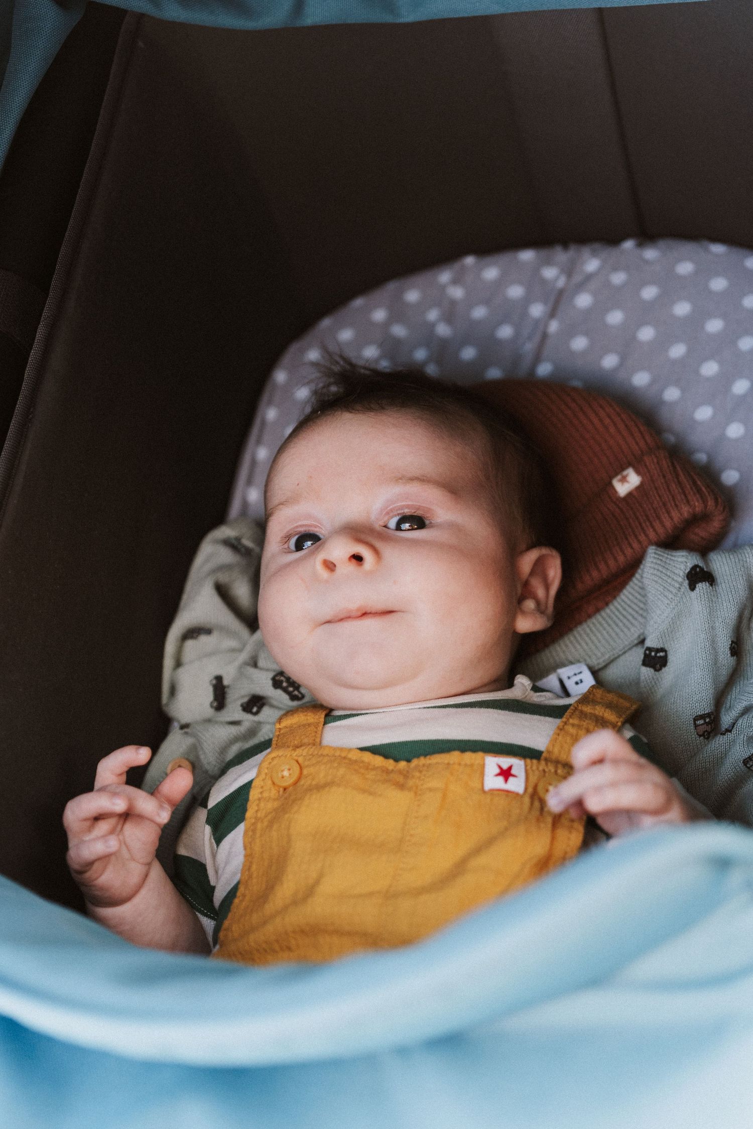 A baby wearing yellow overalls and a brown beanie lies in a polka dot car seat with a light blue blanket.