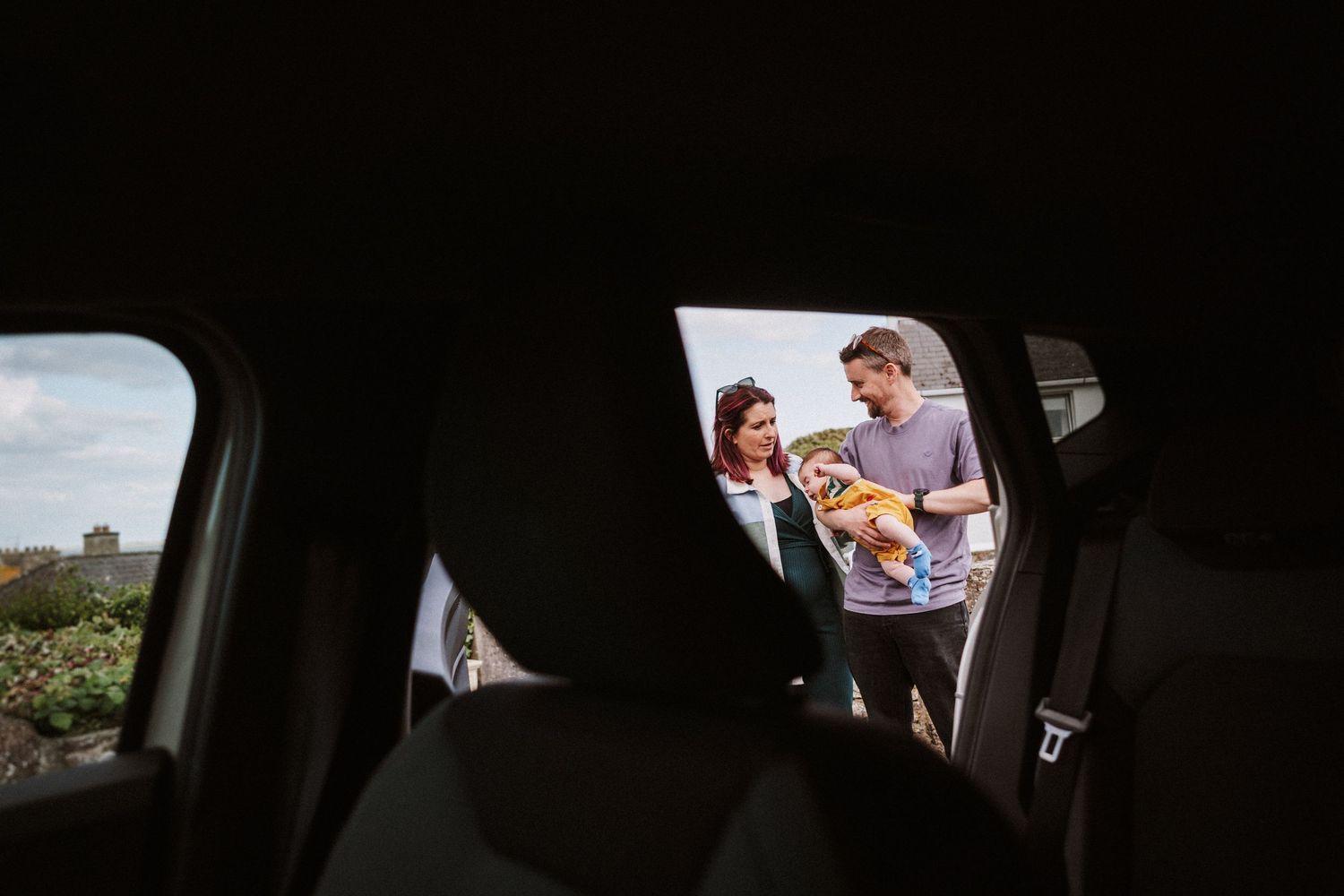 View through car window of a couple standing outside with their baby, framed by the vehicle's interior.