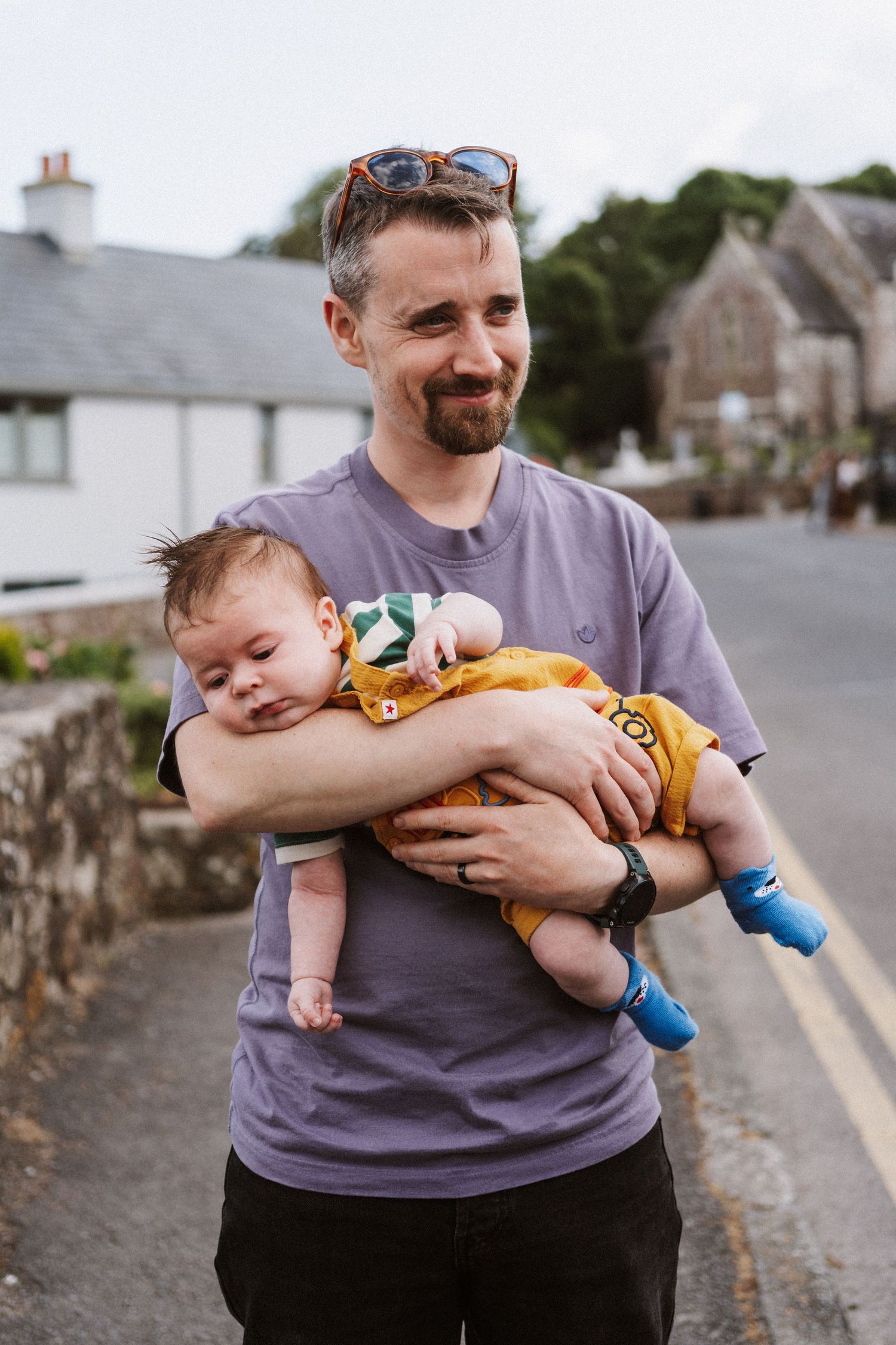 A parent holds a sleepy baby wearing yellow shorts and blue socks on a residential street with stone walls.