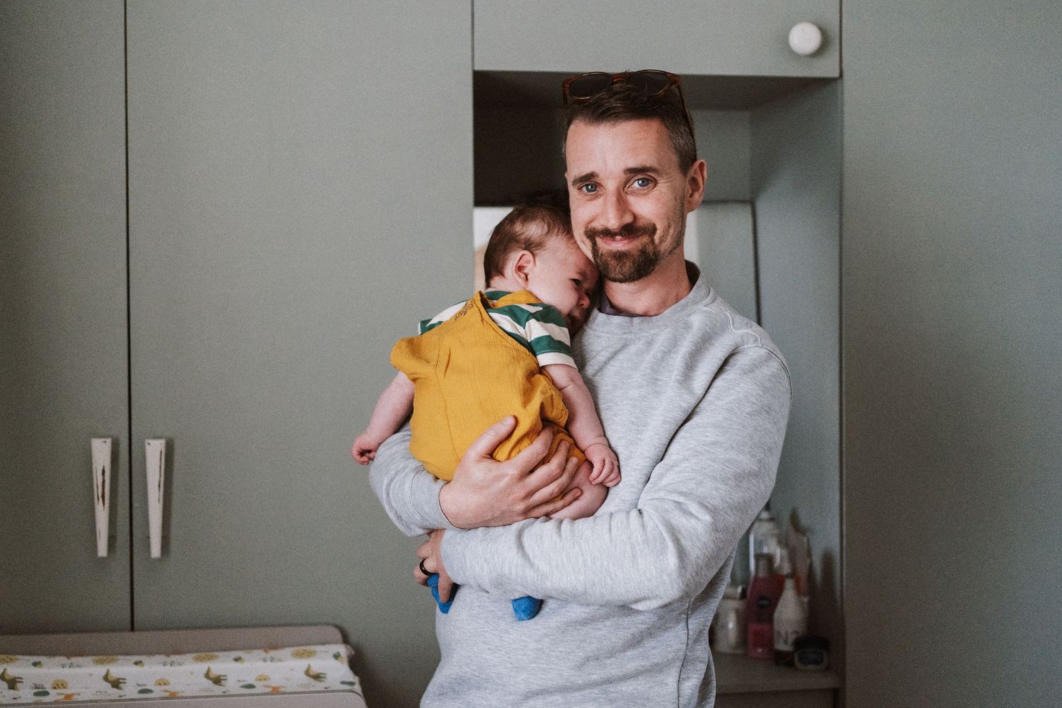A parent in a light colored shirt holds a baby while standing in front of grey cabinets in a kitchen.