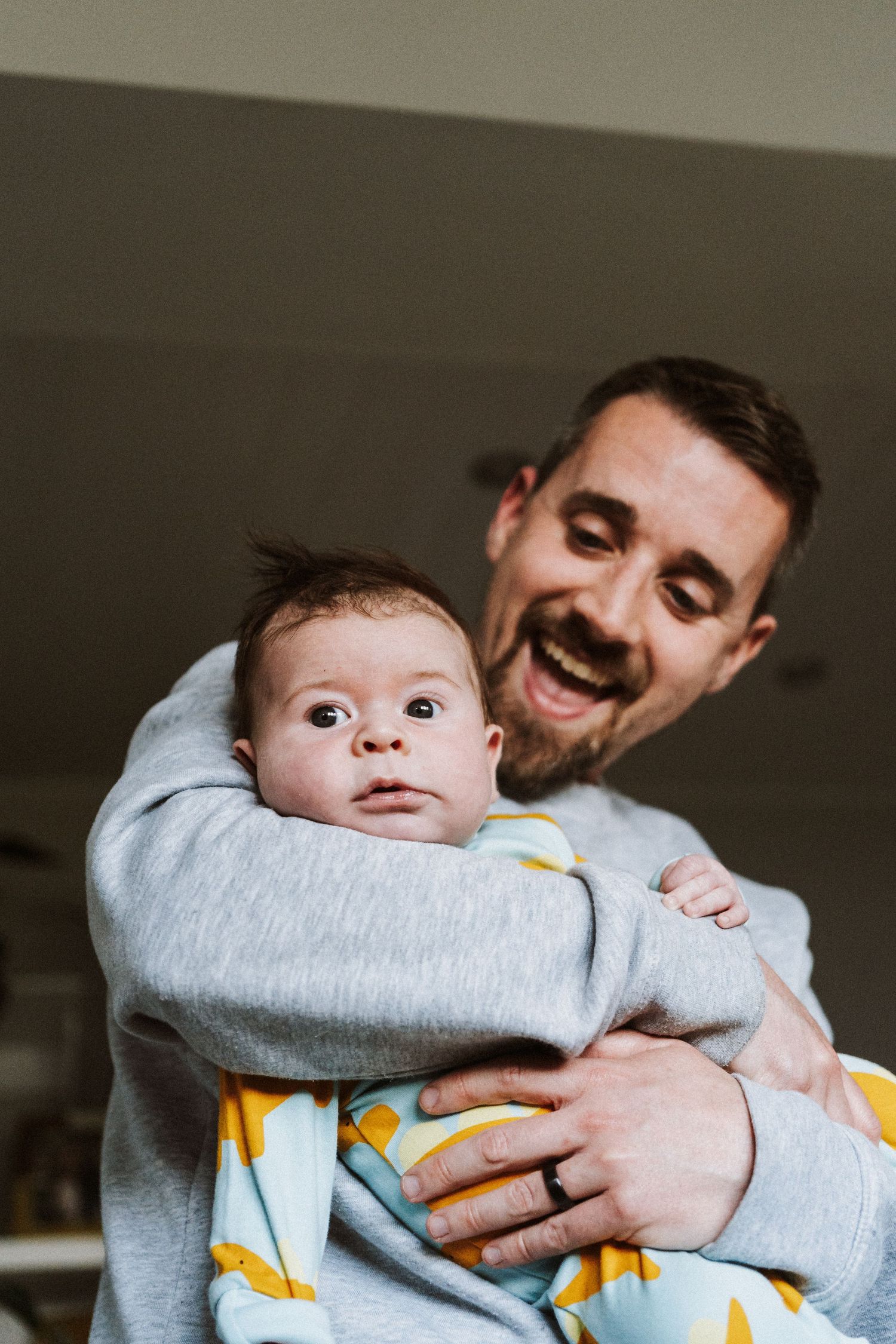 A happy parent holds a wide-eyed baby wearing a gray sweatshirt and yellow patterned blanket.