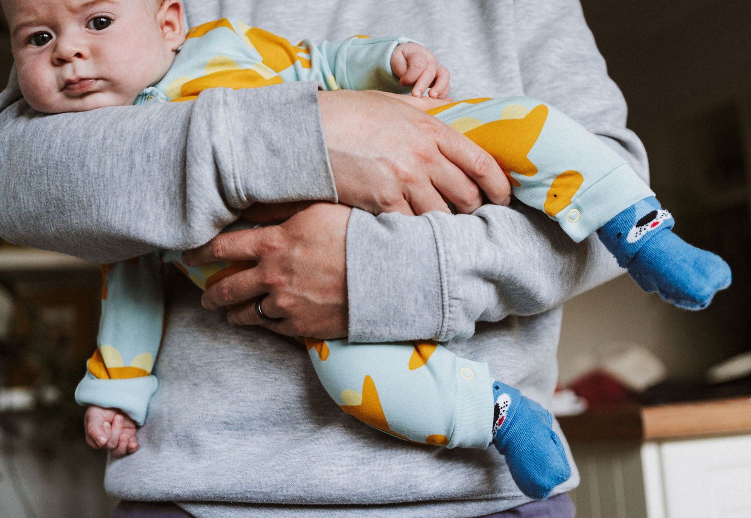 Close-up view of someone holding a baby wearing a yellow and blue patterned outfit against a grey background.