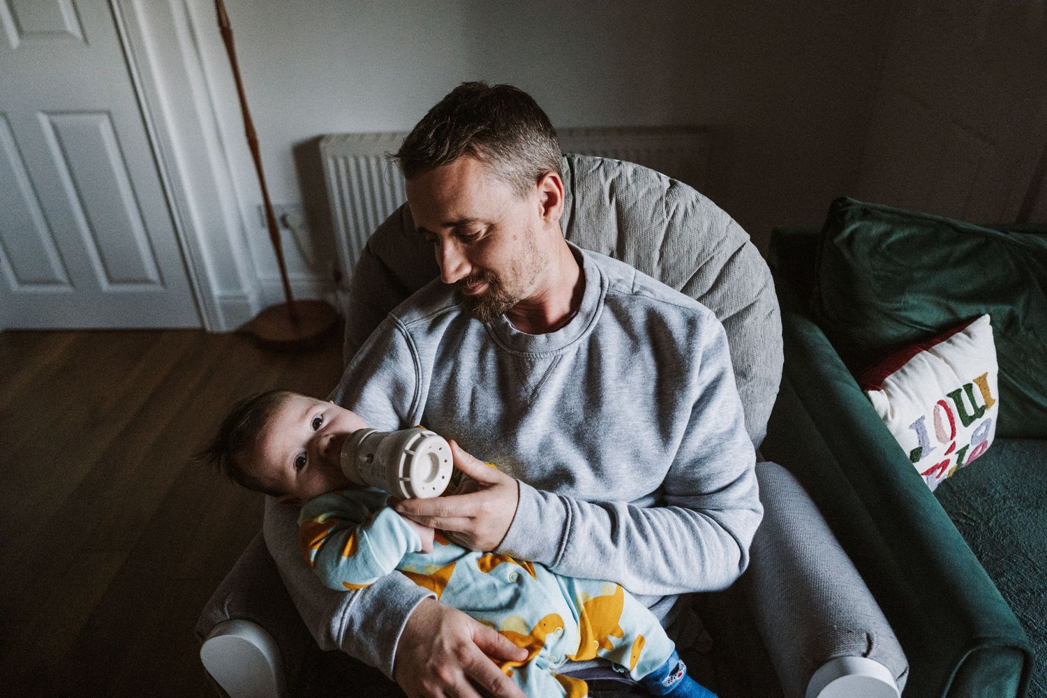 Parent in grey sweatshirt holding and feeding a newborn baby while sitting on a dark green couch.