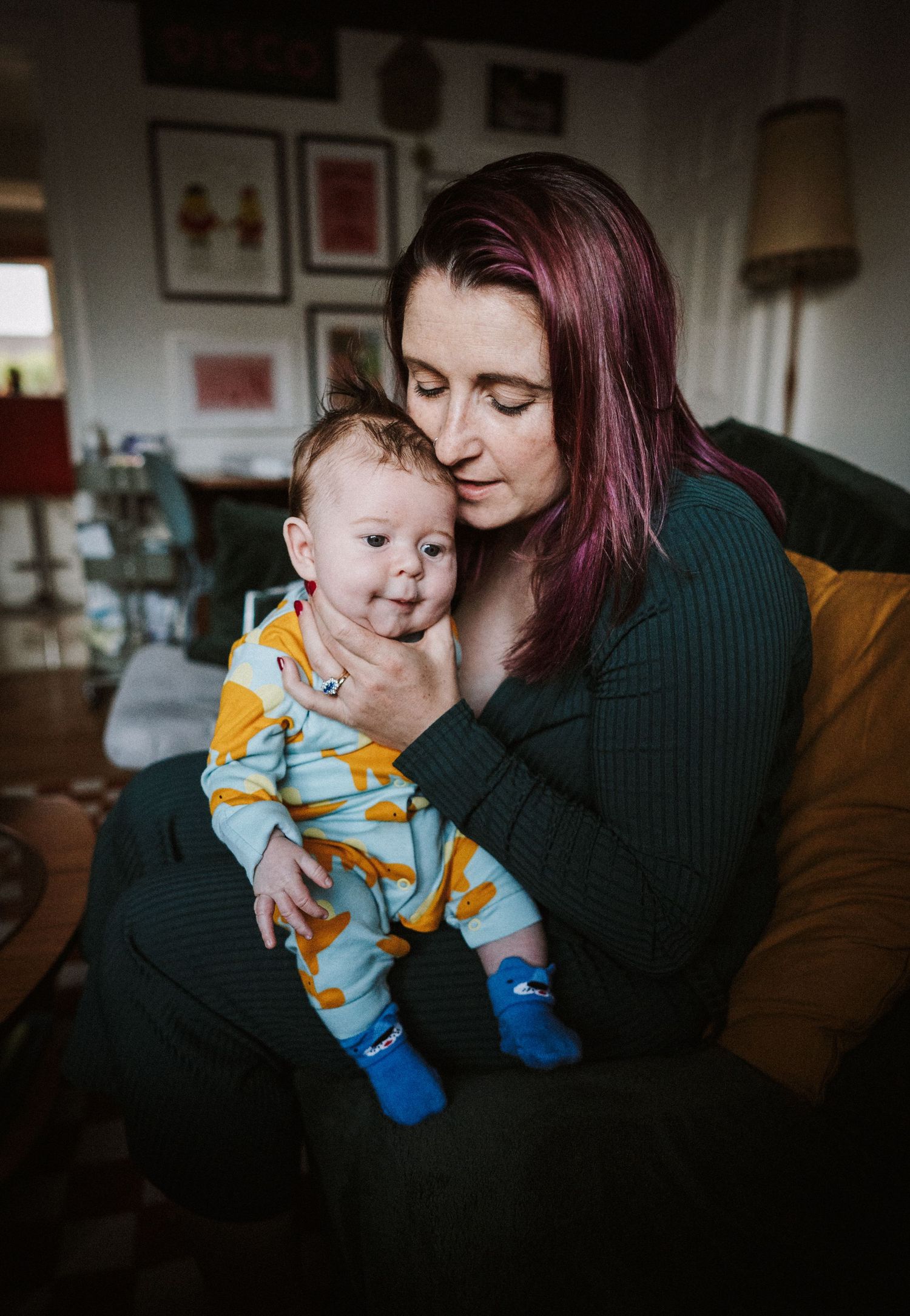 A parent and baby share tender moments on a couch in a dimly lit room with soft natural lighting.