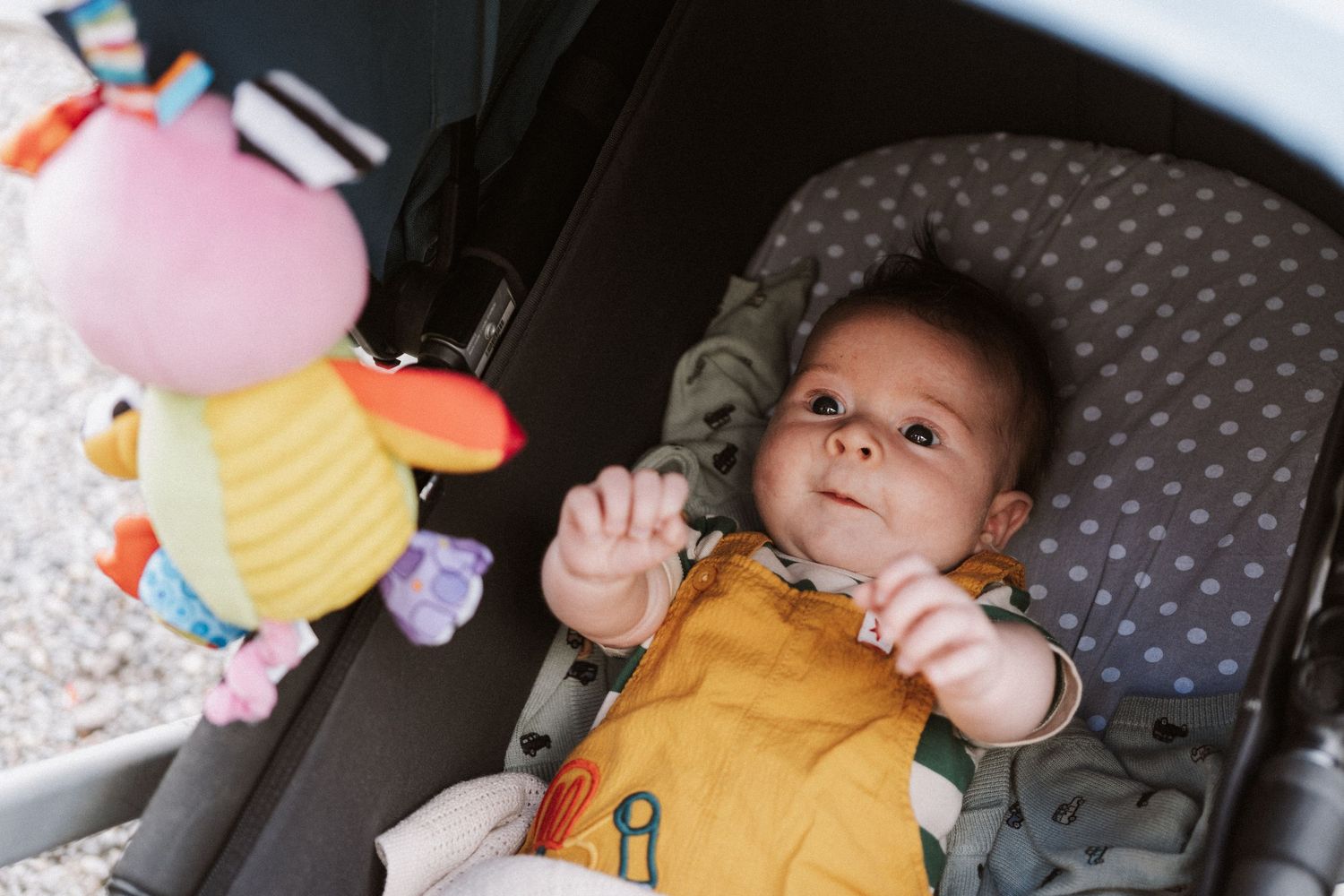A baby in a yellow shirt sits in a car seat reaching up while surrounded by colorful hanging toys and decorations.