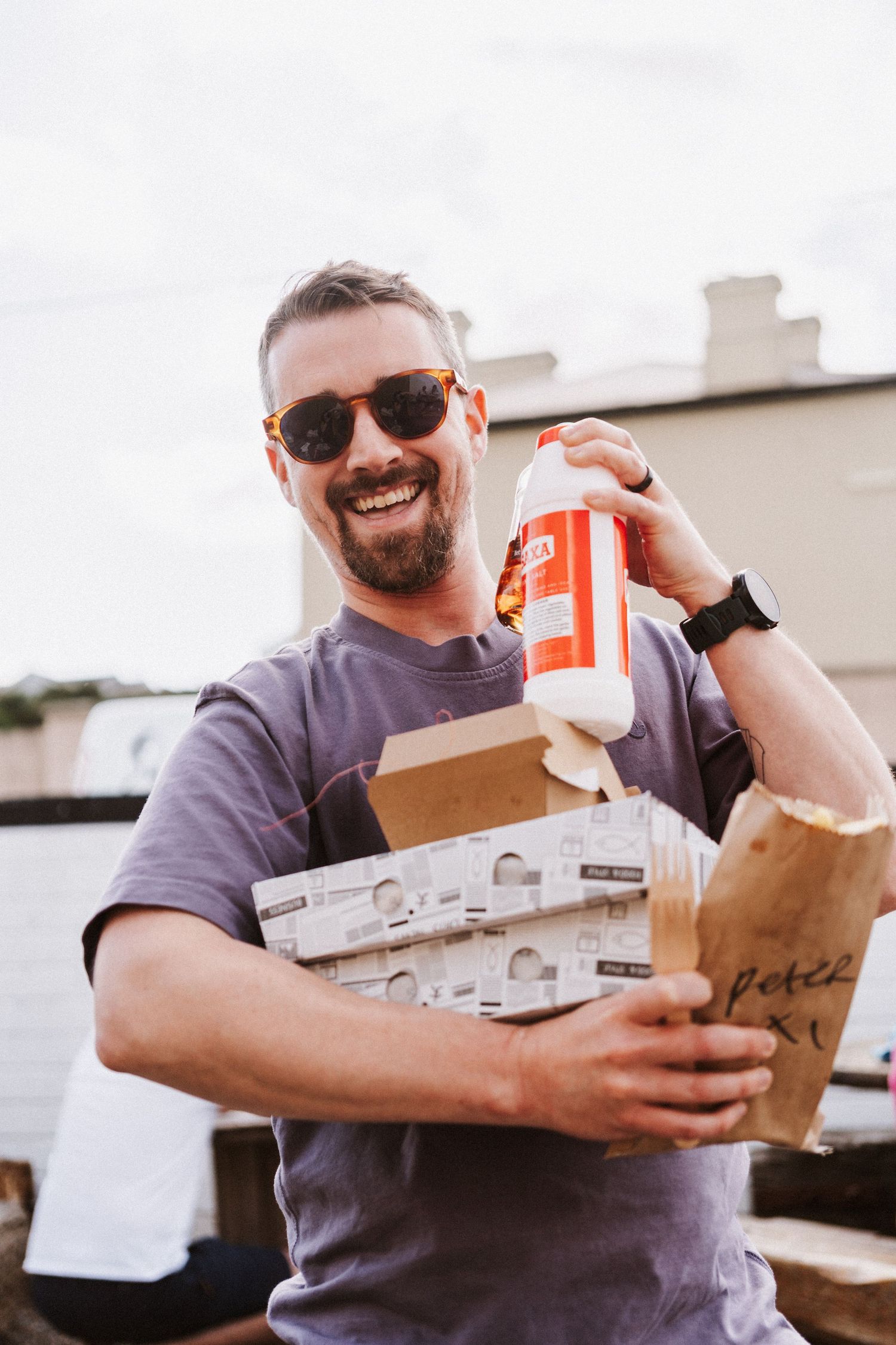 Person in sunglasses and gray shirt joyfully carrying pizza boxes and takeout containers outdoors.
