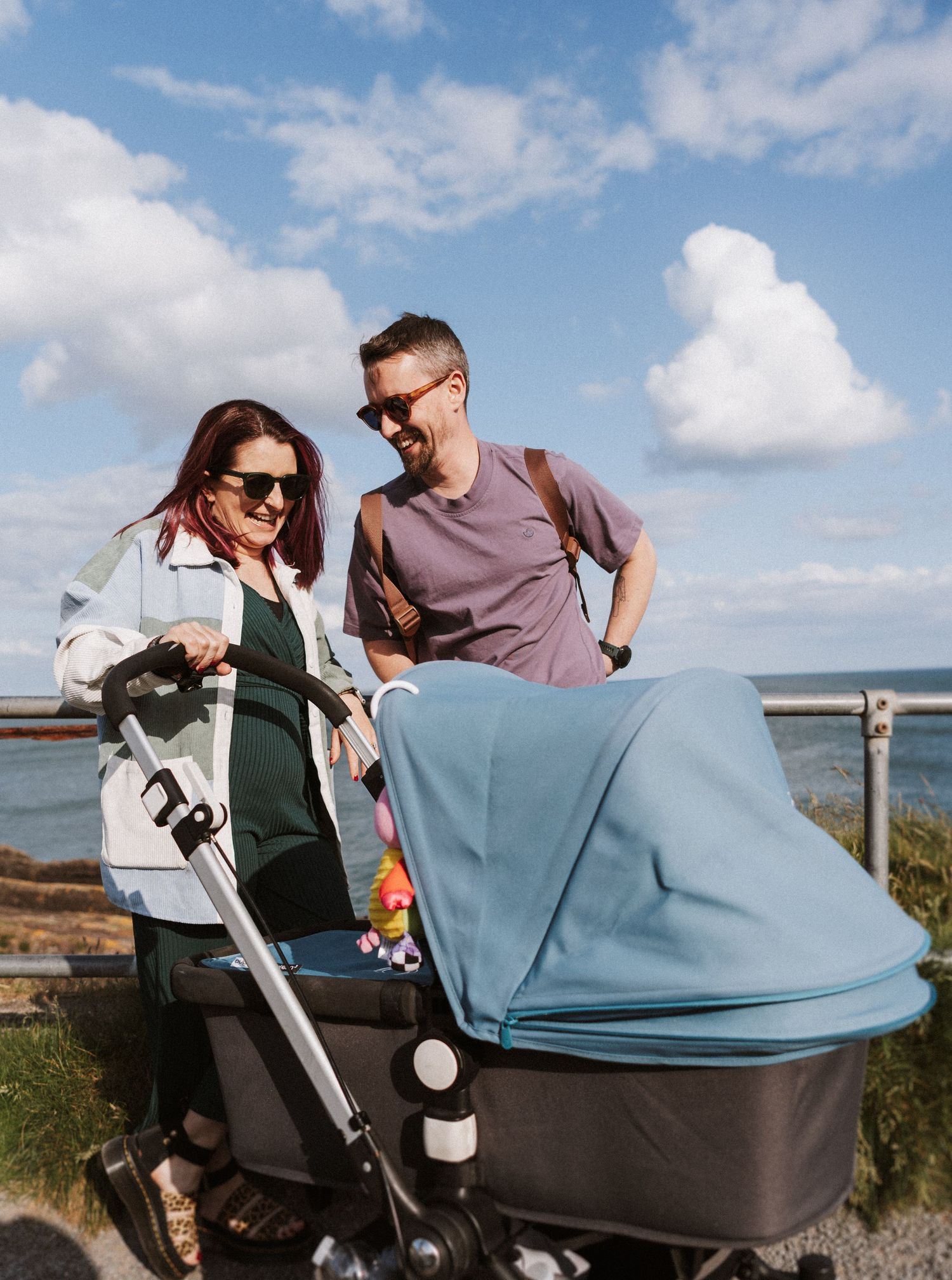 Couple admiring their baby in a stroller during a sunny day at a scenic coastal overlook.