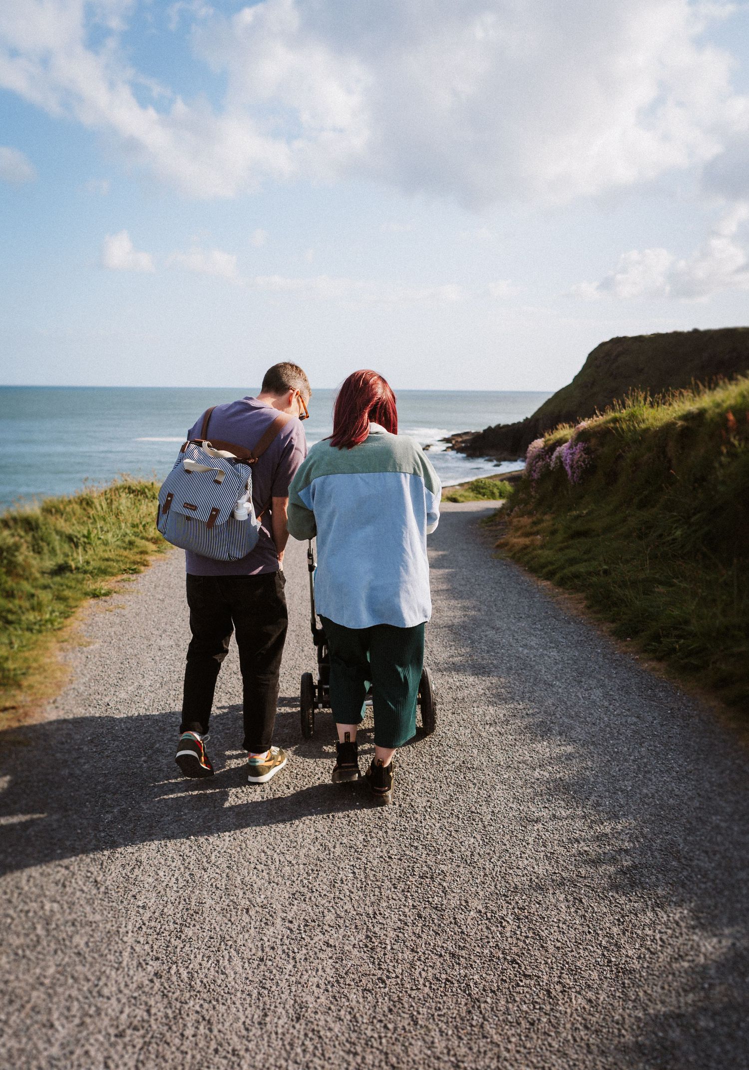 Three people walking together on a scenic coastal path with gravel surface and ocean views on a cloudy day.