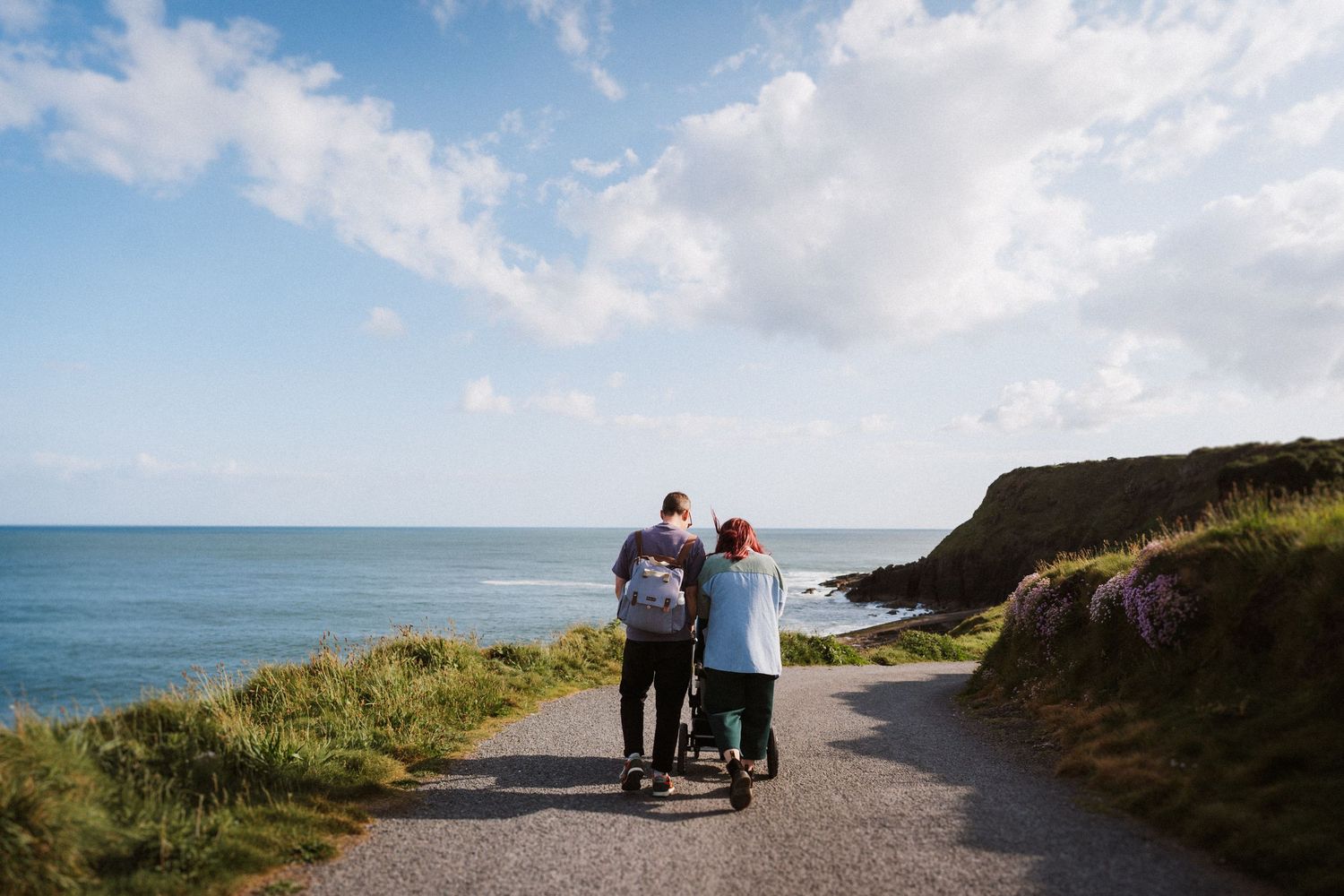 Three people walk together on a scenic coastal path overlooking the ocean on a sunny day with blue skies and clouds.