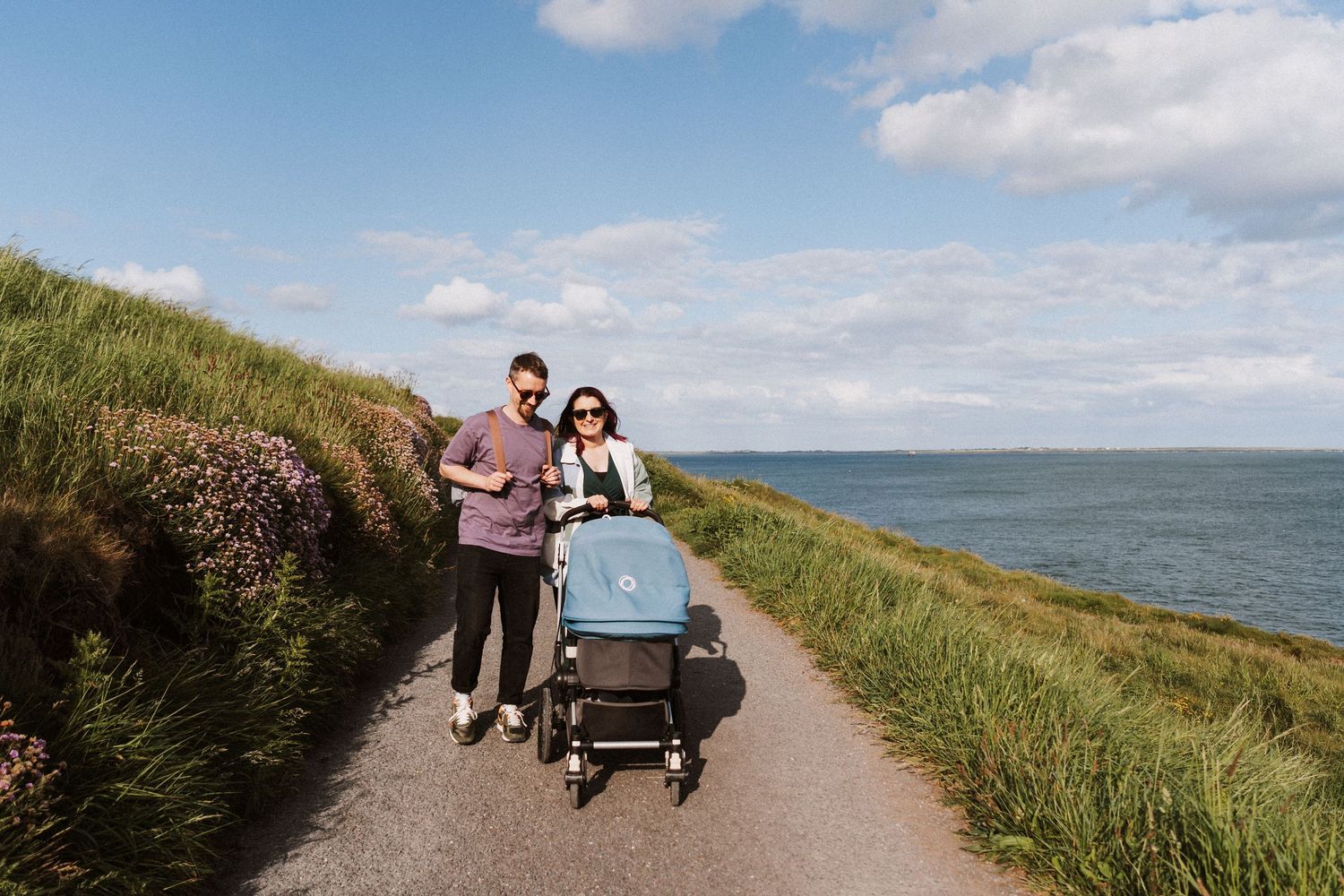 A couple walks with a stroller along a scenic coastal path with grassy cliffs and ocean views on a sunny day.