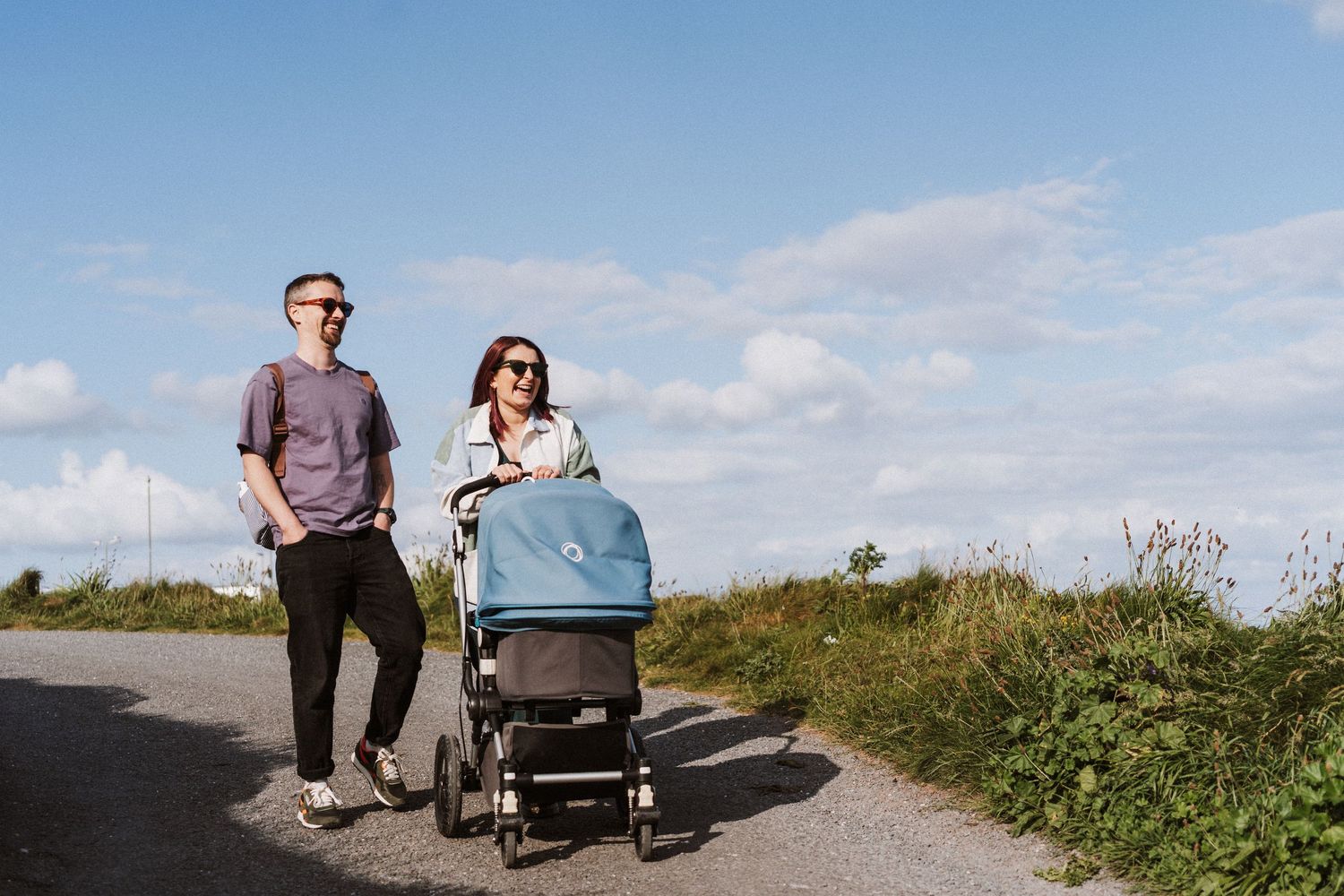 People walking with stroller on a scenic coastal path with blue sky and wildflowers on a sunny day.