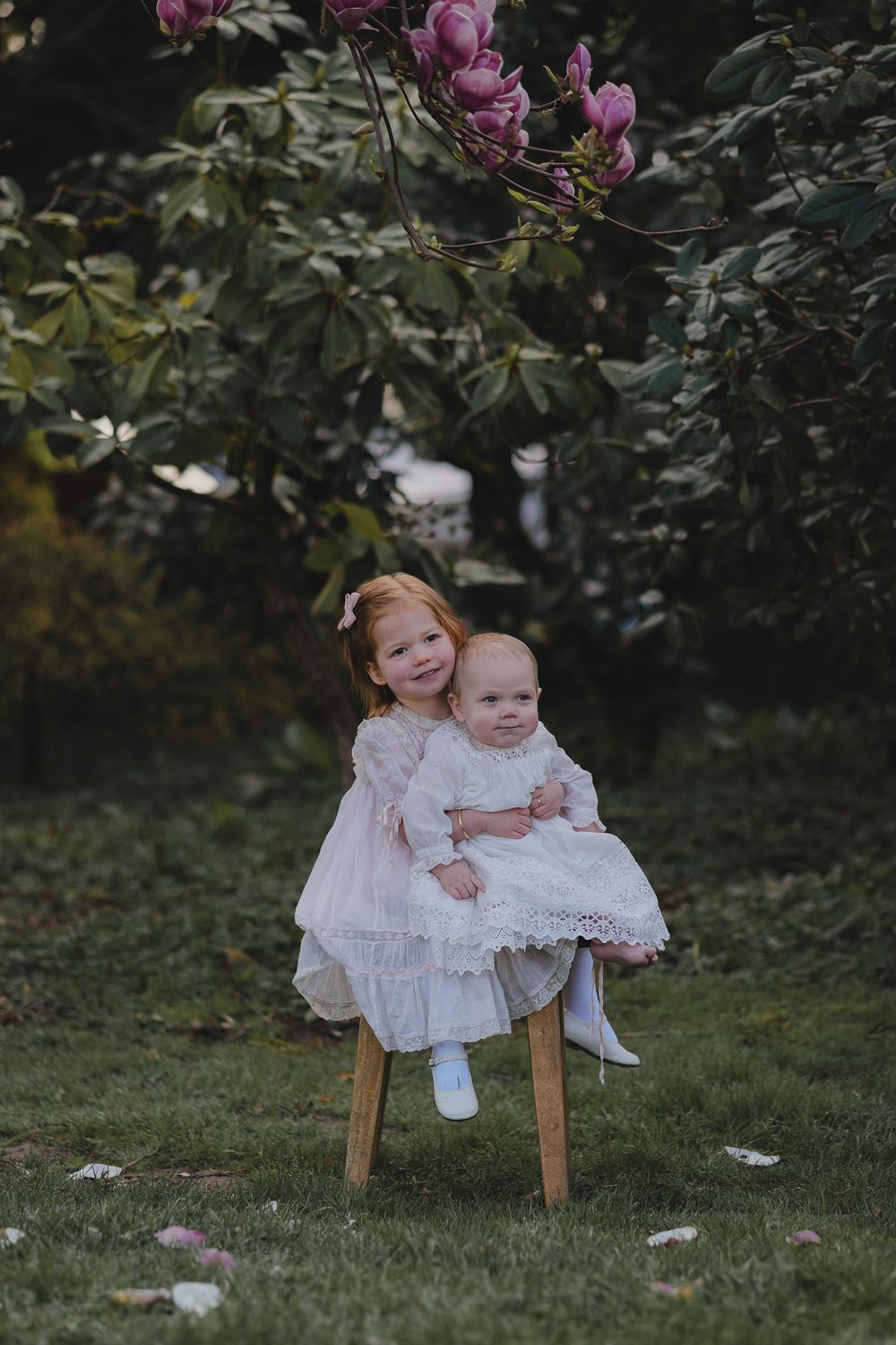 Two children in white dresses sit together on wooden chairs in a garden setting with green foliage in the background.