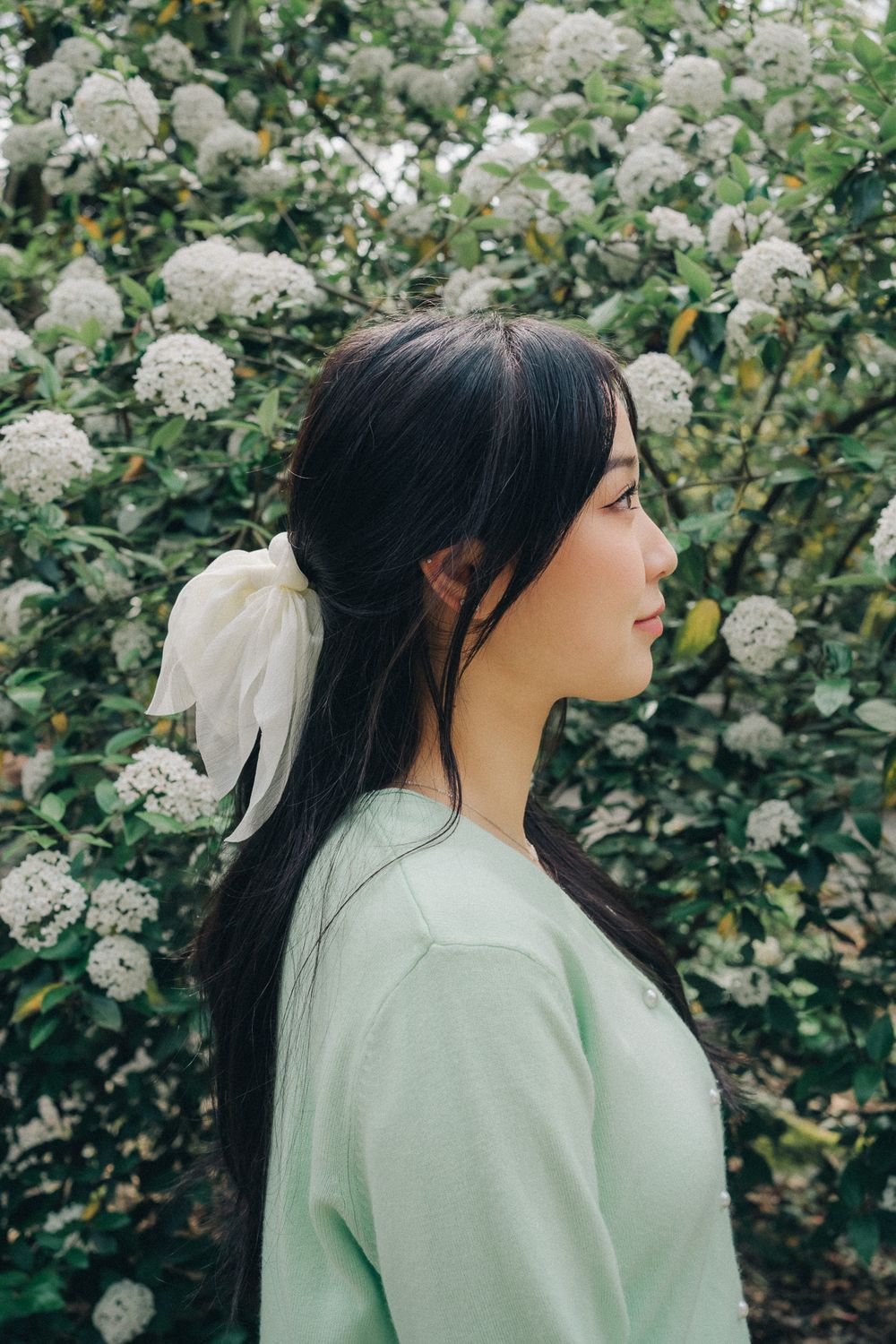Side profile portrait in a mint green sweater against white flowering bushes in a garden setting.