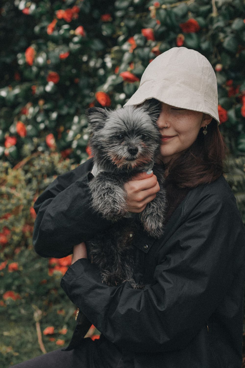 A person in a black jacket and beige cap shares tender moments with a small gray dog in a garden setting.
