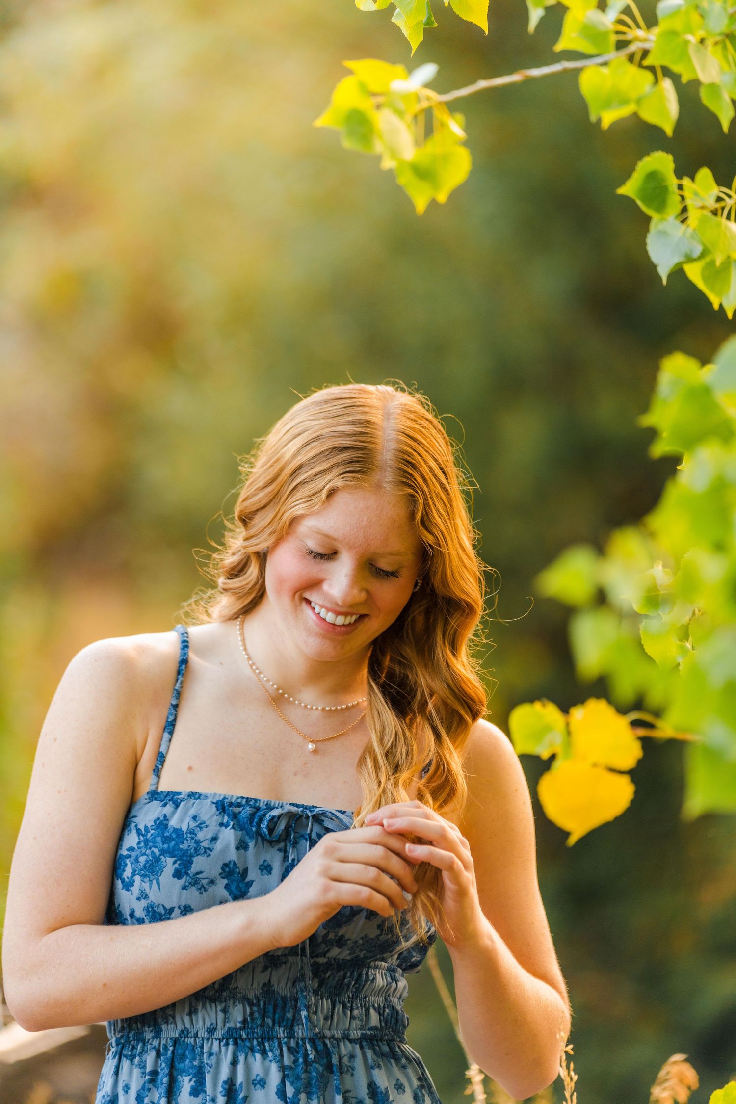 Smiling redhead teen in a blue floral dress stands under green leaves in warm golden light.