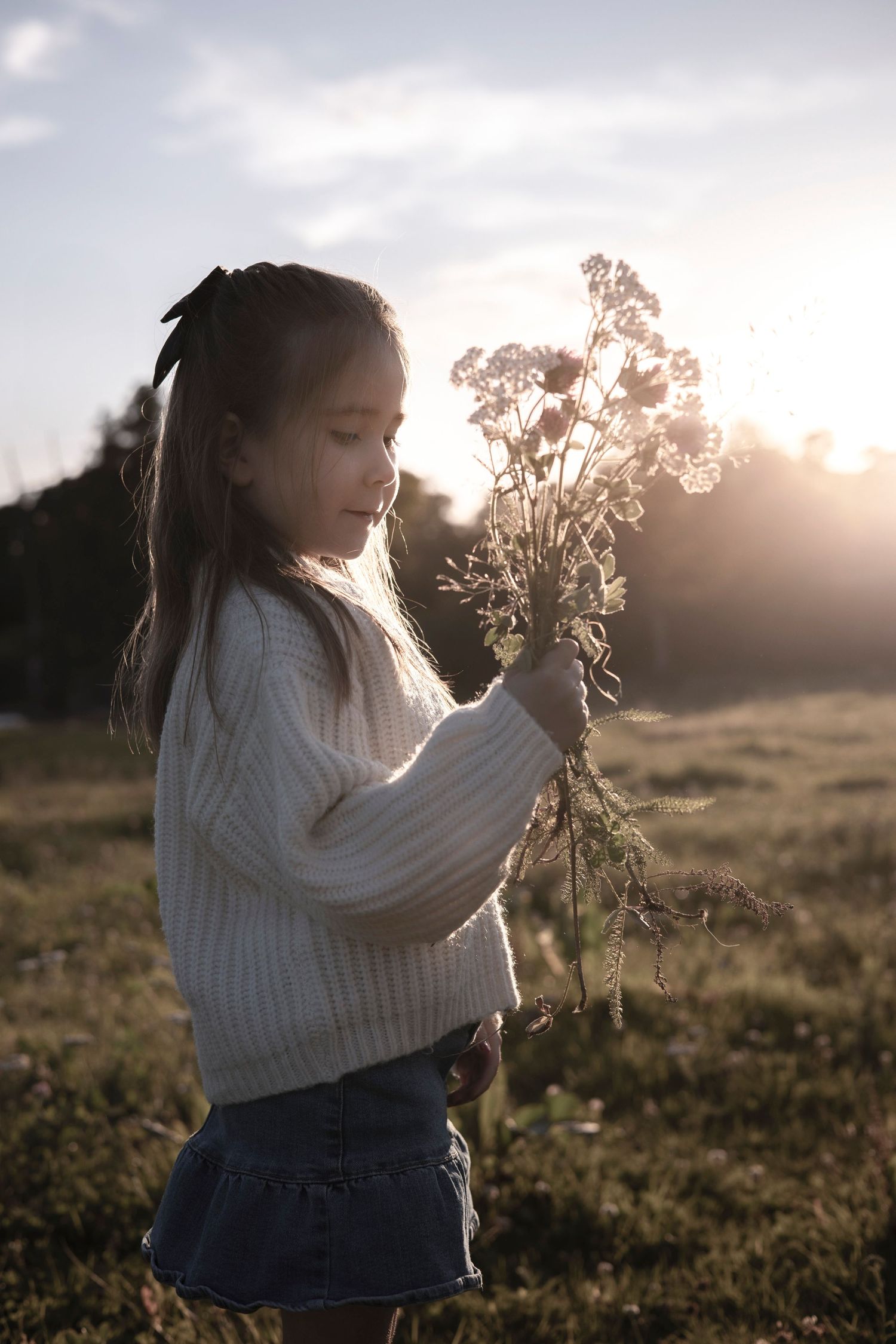 flicka står på en äng och håller i en blombukett i bakgrunden strålar solen