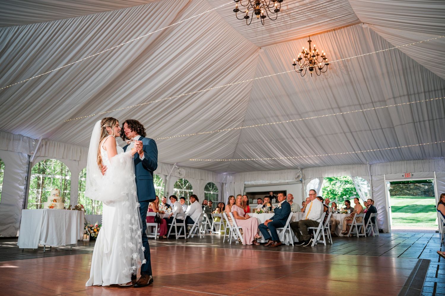A romantic dance under a white draped tent ceiling with elegant chandeliers at an outdoor wedding reception.
