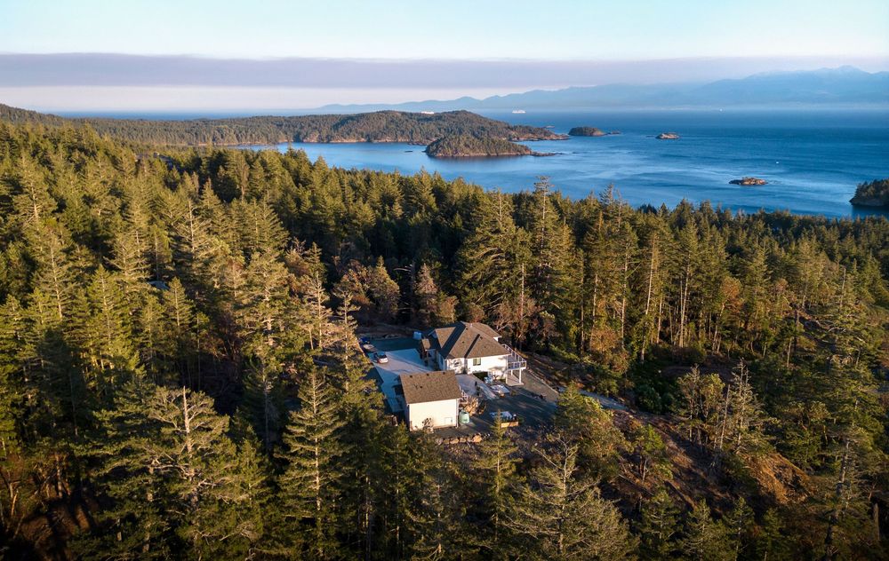 Aerial view of a secluded house nestled in dense forest overlooking a scenic ocean bay.