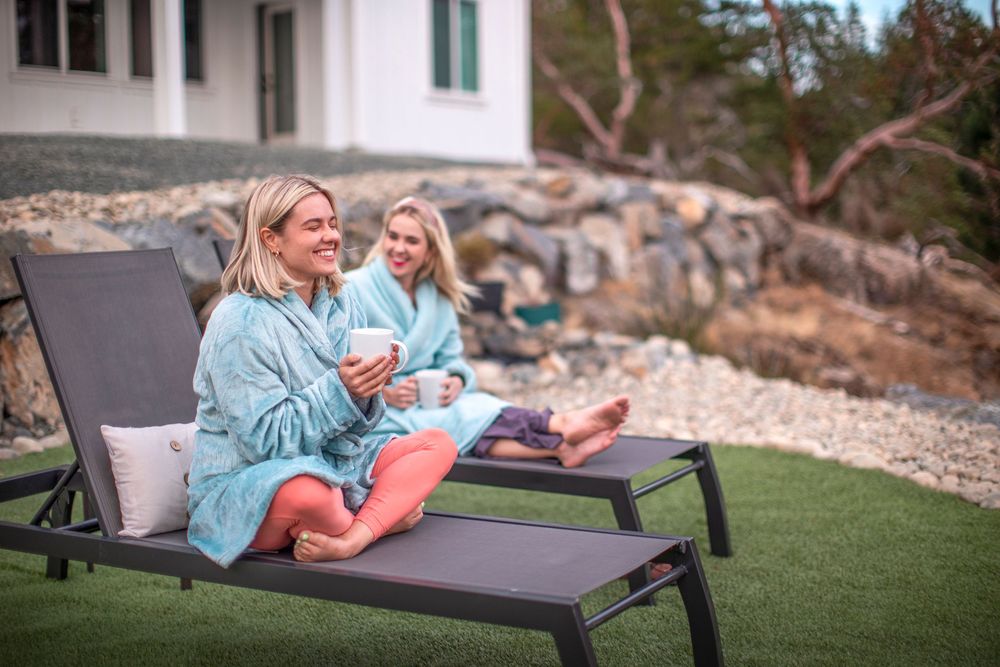 Two blonde women relaxing on lounge chairs in robes outdoors with ocean view backdrop.