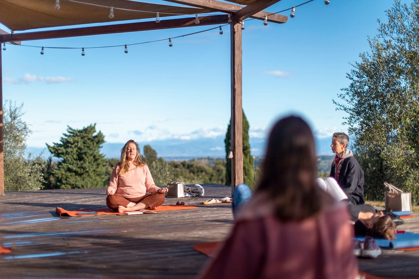 Outdoor yoga class on wooden deck with instructor meditating in lotus pose under pergola with scenic sky view.