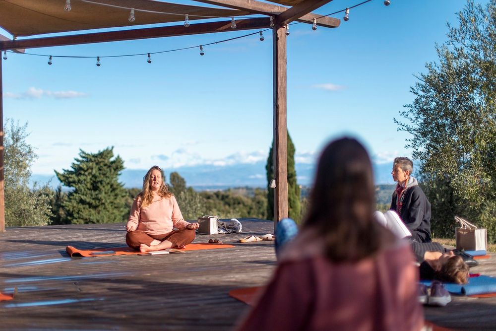 Outdoor yoga class on wooden deck with instructor meditating in lotus pose under pergola with scenic sky view.