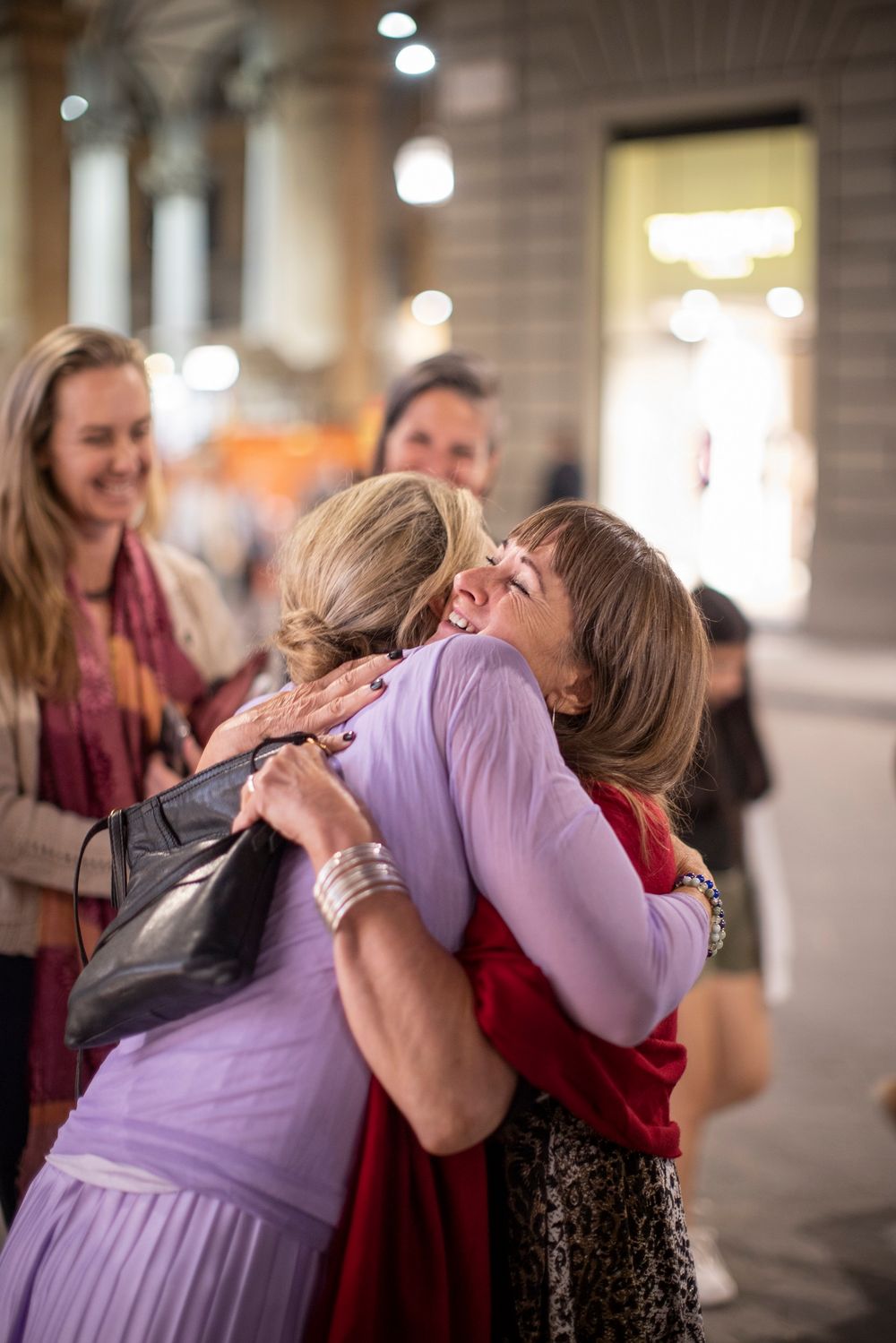 Two women share a warm embrace outdoors, smiling joyfully, with other women socializing in the blurred background.