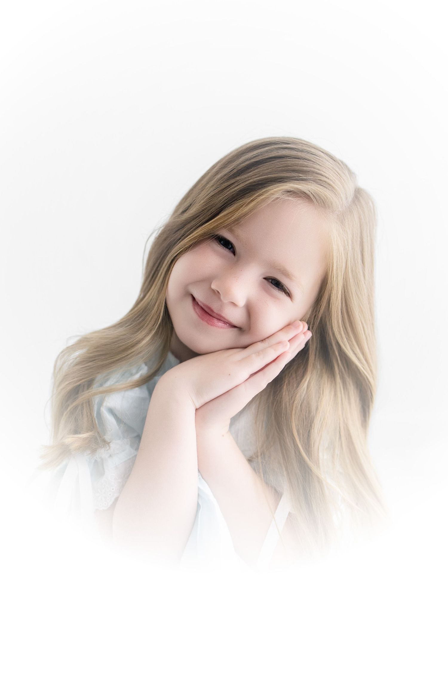 A smiling young girl with long blonde hair rests her hands on her cheek against a soft white background.