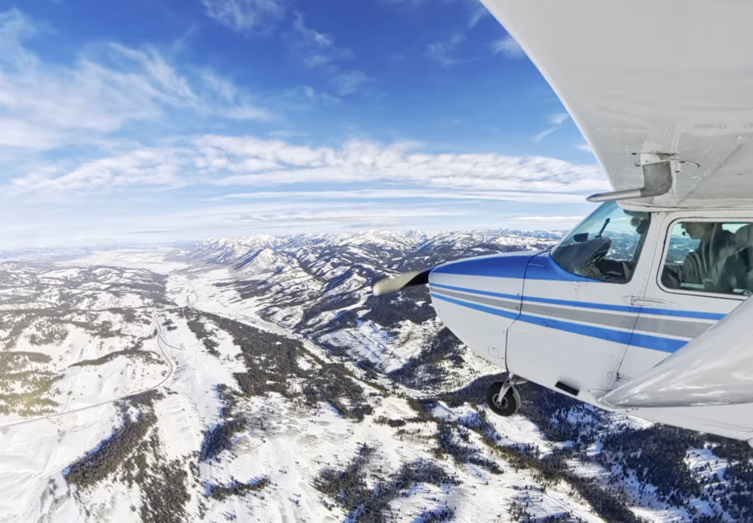 Small blue and white plane flying over snow-capped mountains under a bright blue sky. Captured by Slate Media (best in UT)
