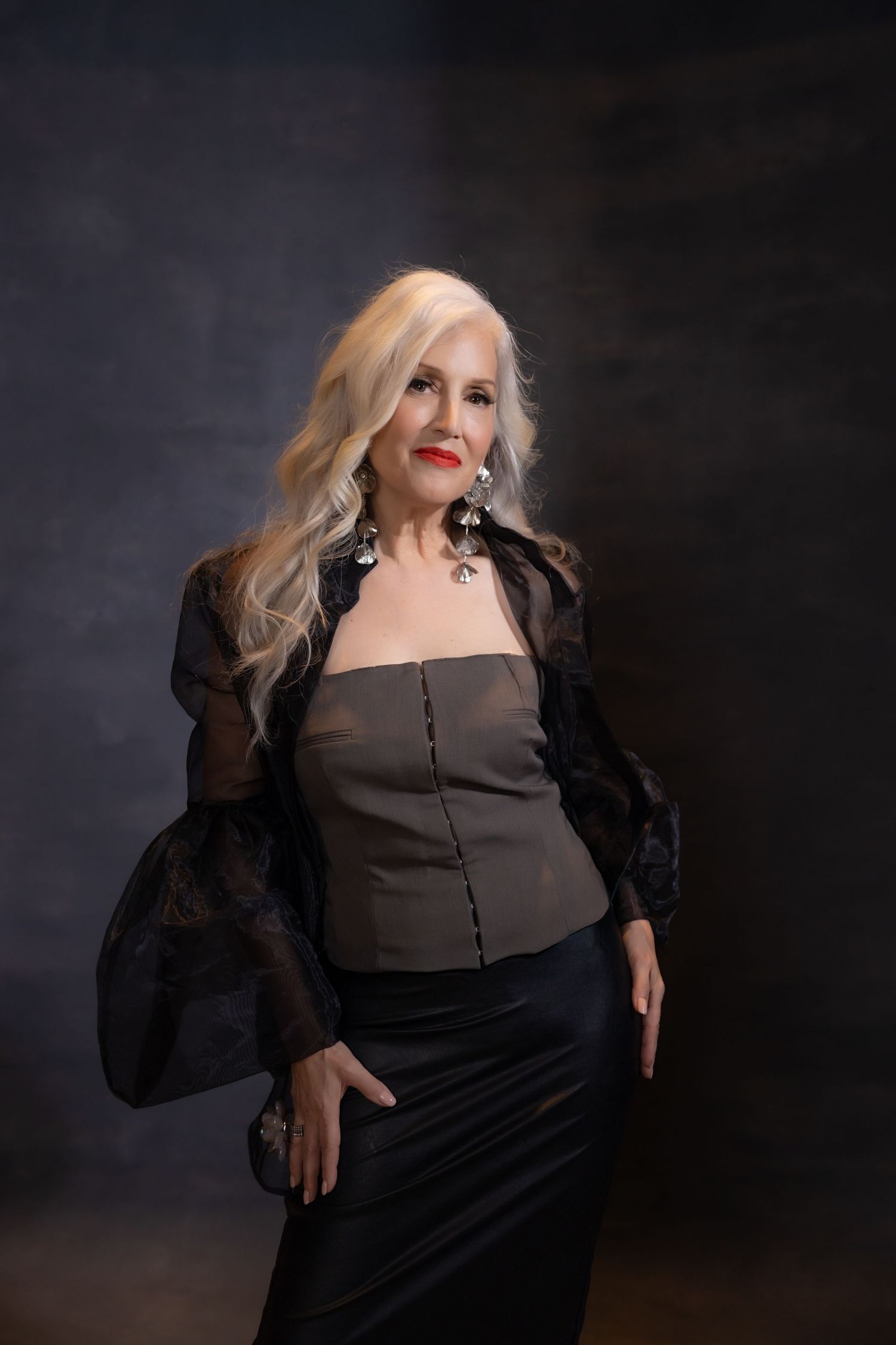 Elegant mature blonde woman in black outfit with red lips posing confidently against dark studio backdrop.