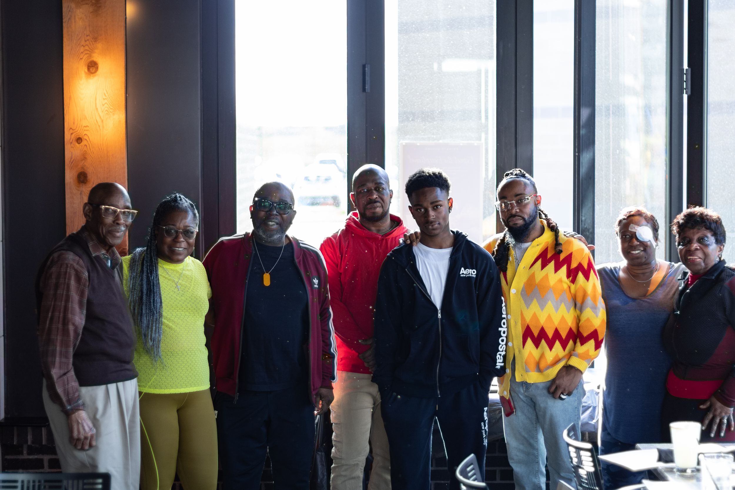 Eight people smiling for a group portrait in a sunlit indoor space with large windows and casual, colorful clothing.