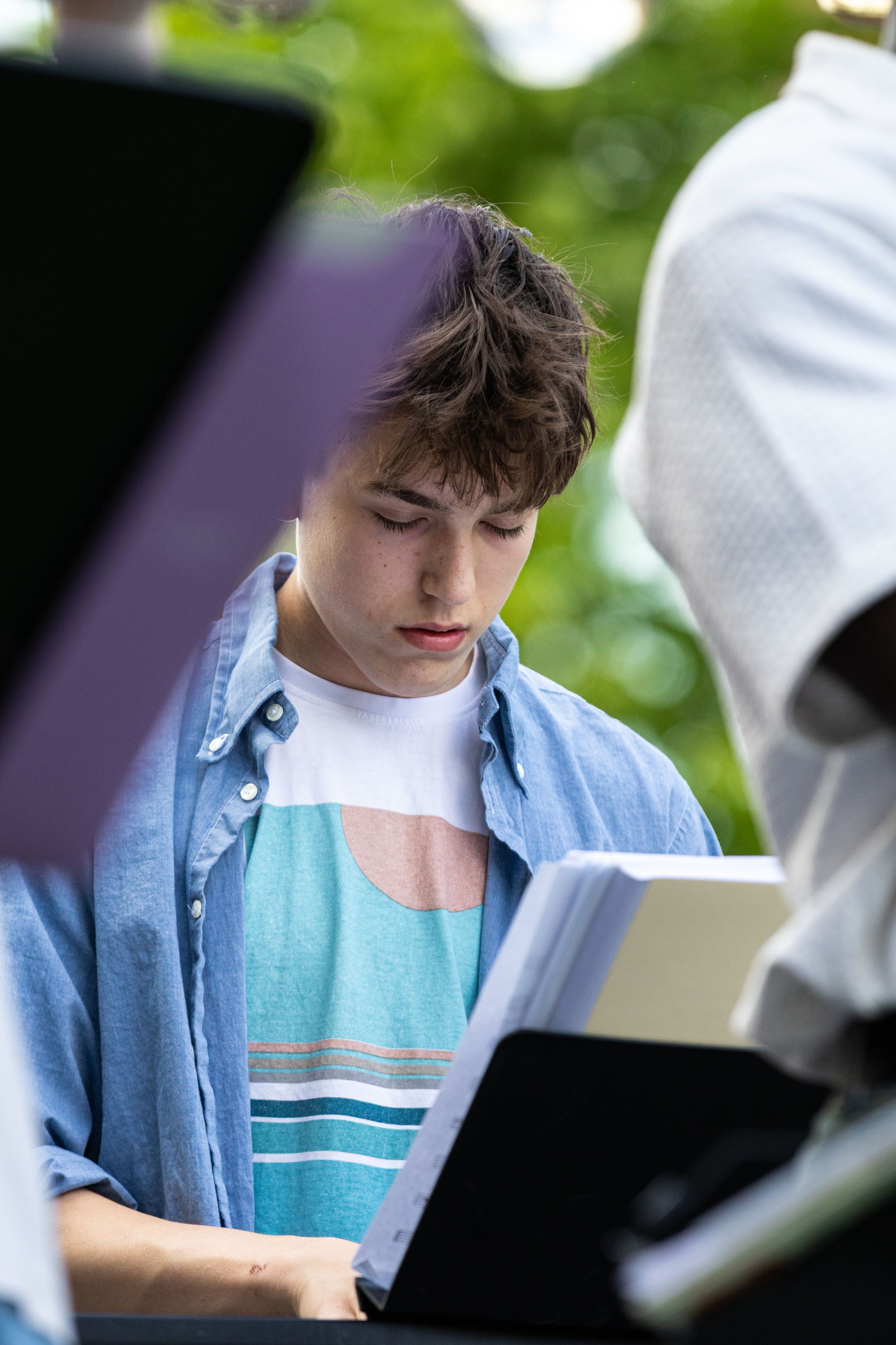 A teenage boy in a blue shirt reads sheet music outdoors at a music stand.